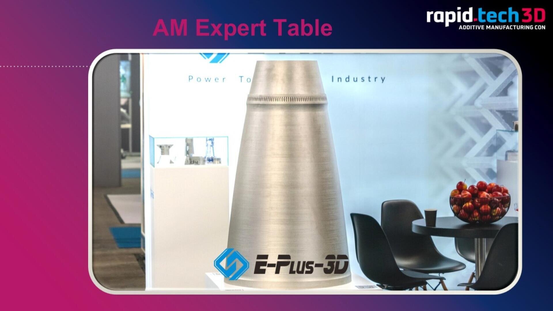 A large metallic 3D-printed cone labeled E-Plus-3D is displayed next to a table with black chairs and a bowl of apples at a trade show booth. The background features branding for an additive manufacturing event.