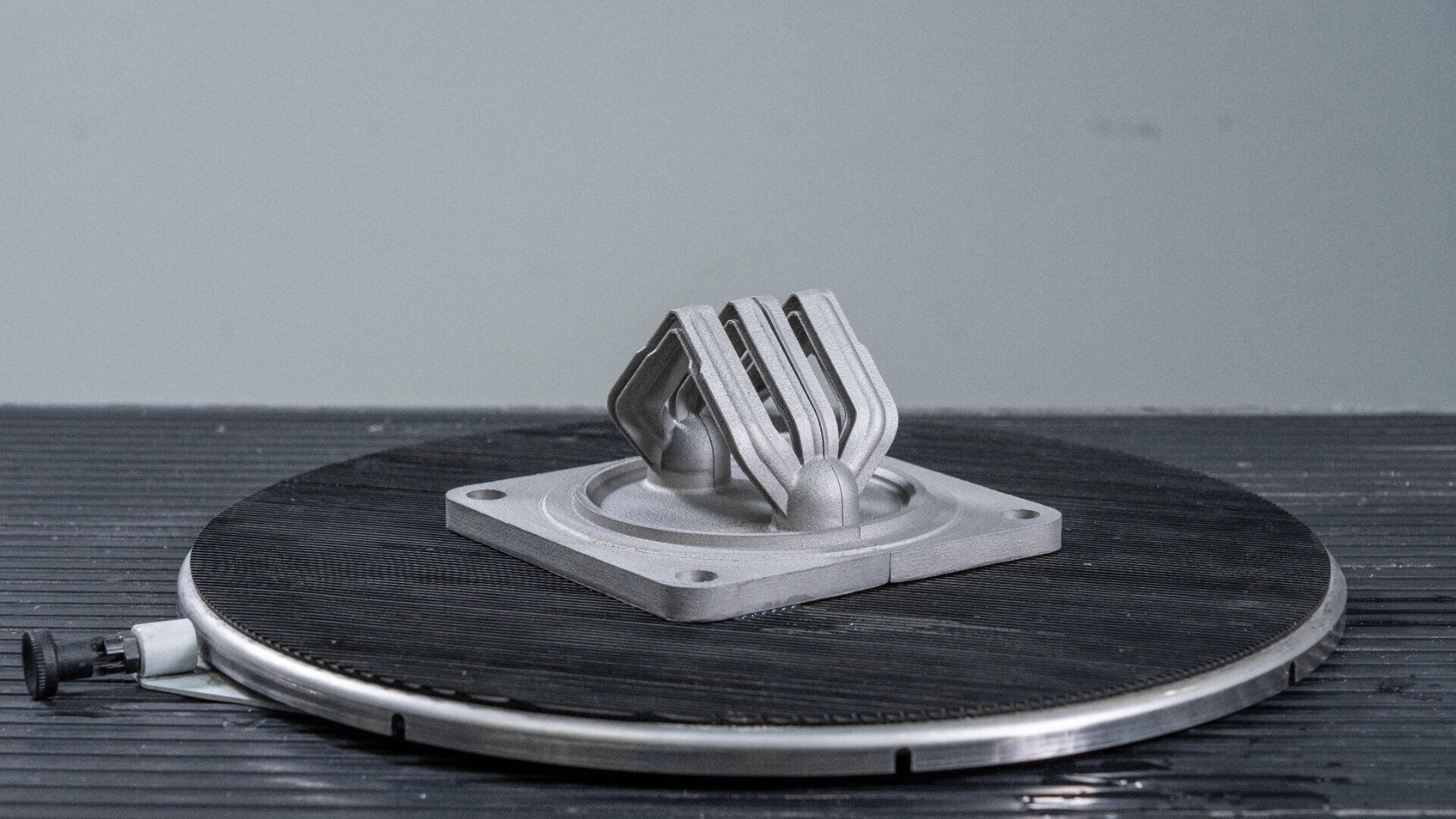 Ein kleines metallisches, 3D-gedrucktes mechanisches Teil mit geometrischen Ausschnitten sitzt auf einer runden schwarzen Plattform vor einem schlichten grauen Hintergrund.