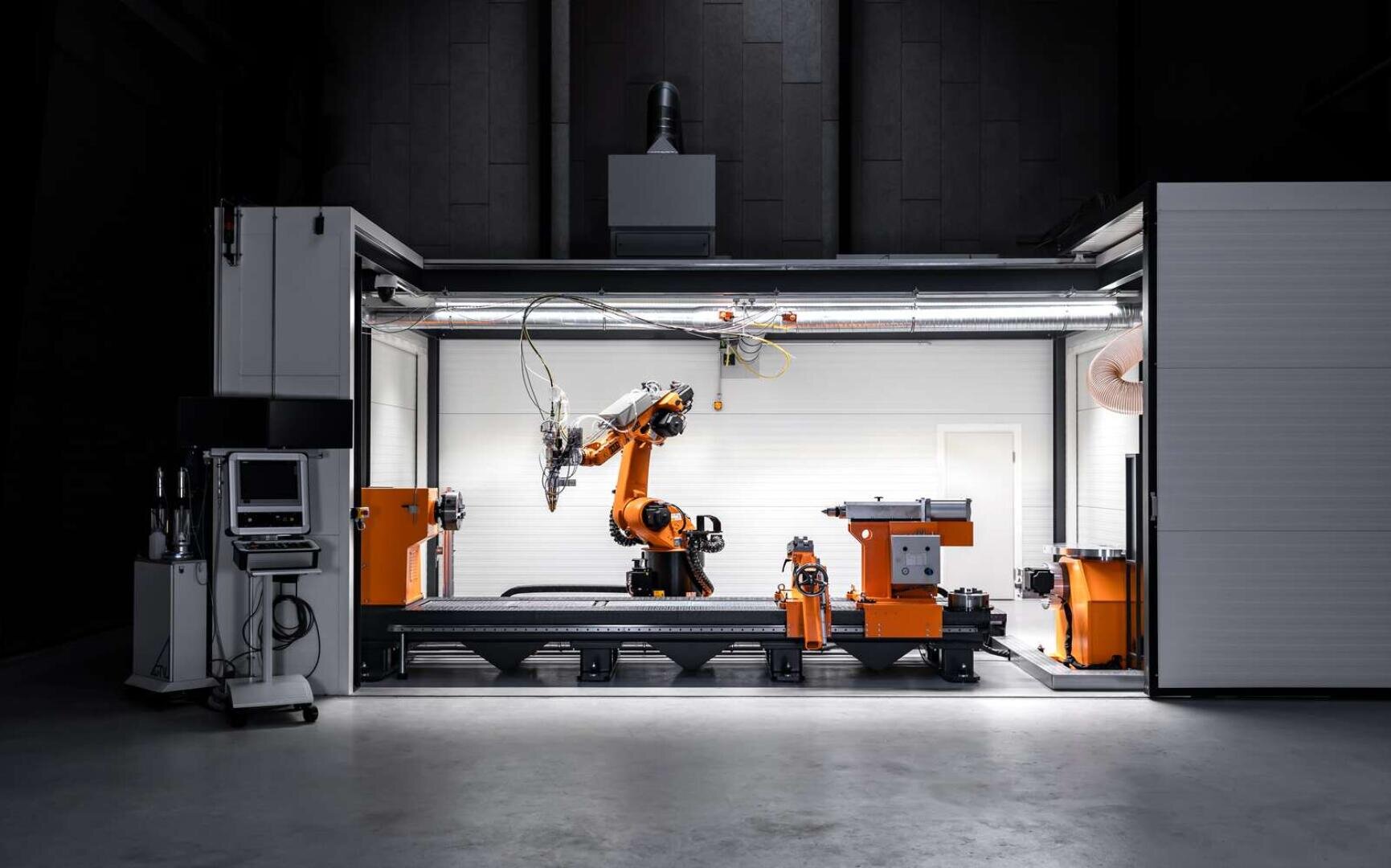 Ein moderner Industrieroboterarm arbeitet in einer gut beleuchteten Fertigungszelle, umgeben von orangefarbenen und weißen Maschinen und Geräten, in einer ansonsten dunklen Fabrikumgebung.