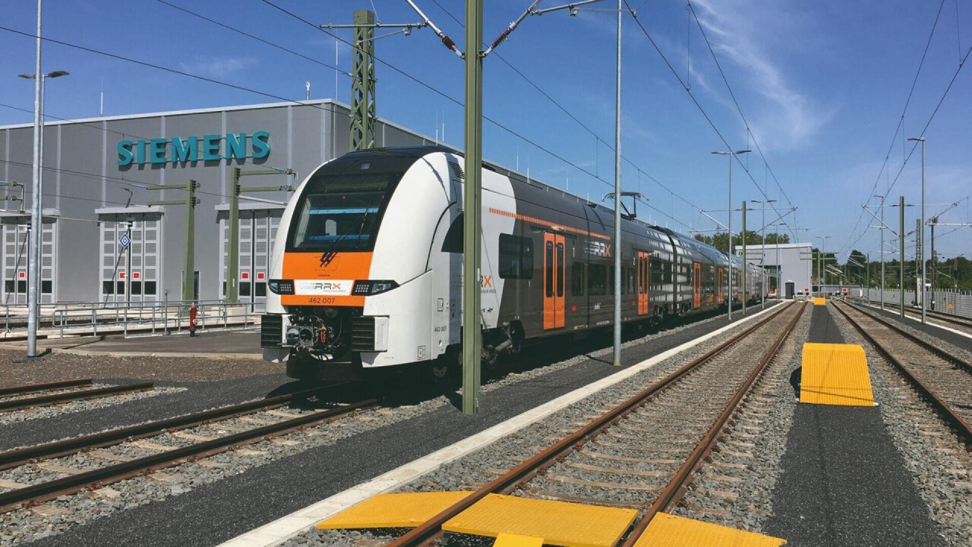 A modern white and orange passenger train is on railway tracks outside a Siemens building on a clear, sunny day. Overhead electric wires and multiple tracks are visible in the scene.