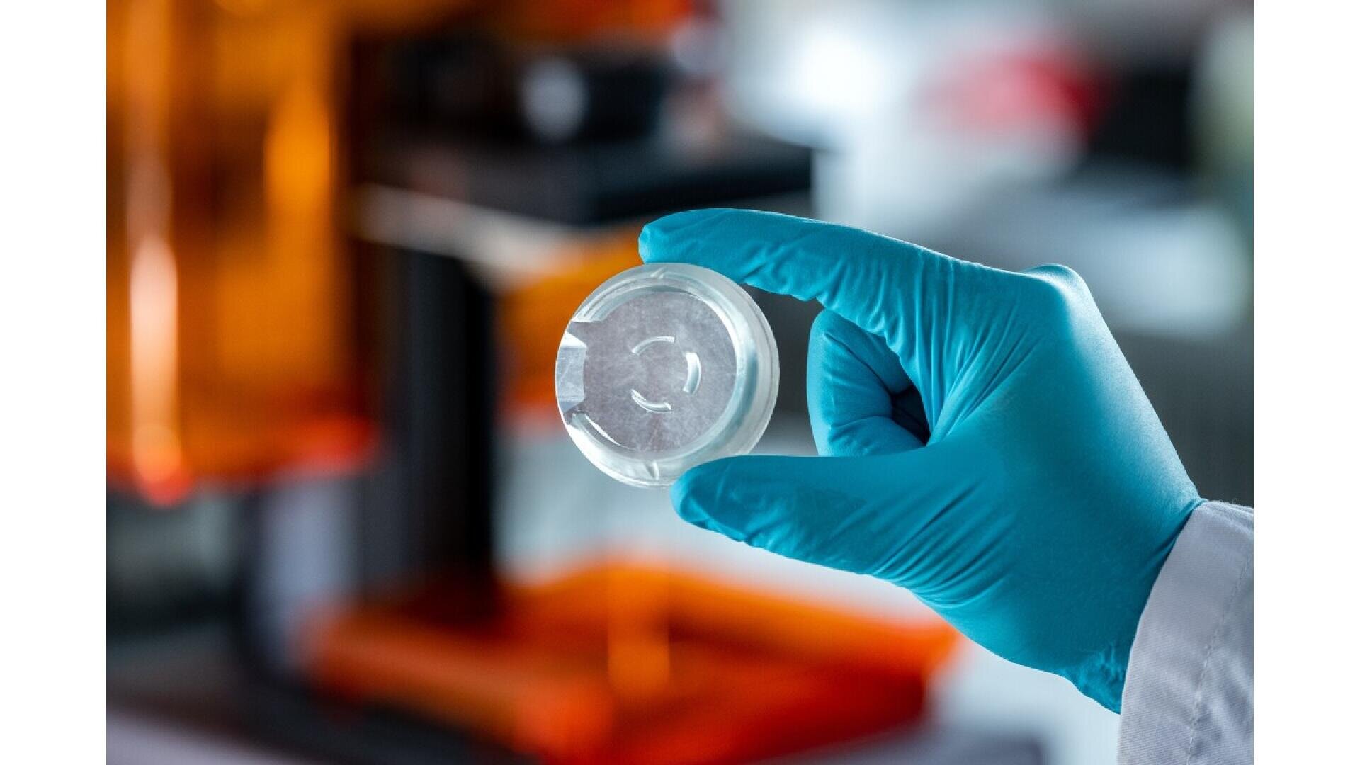 A gloved hand holds up a clear petri dish with a sample inside, in a laboratory setting with scientific equipment visible in the blurred background.