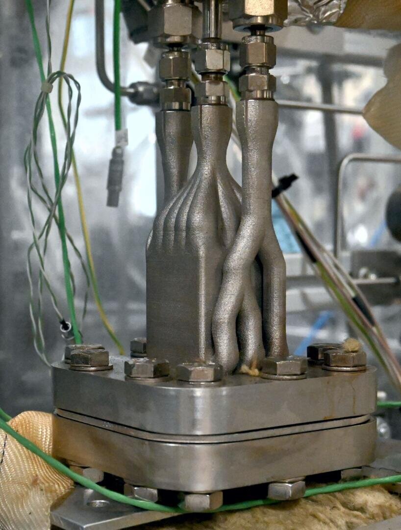 Close-up of a metallic, 3D-printed rocket engine part with twisted, tubular channels and bolted connections, surrounded by electrical wires and industrial equipment in the background.