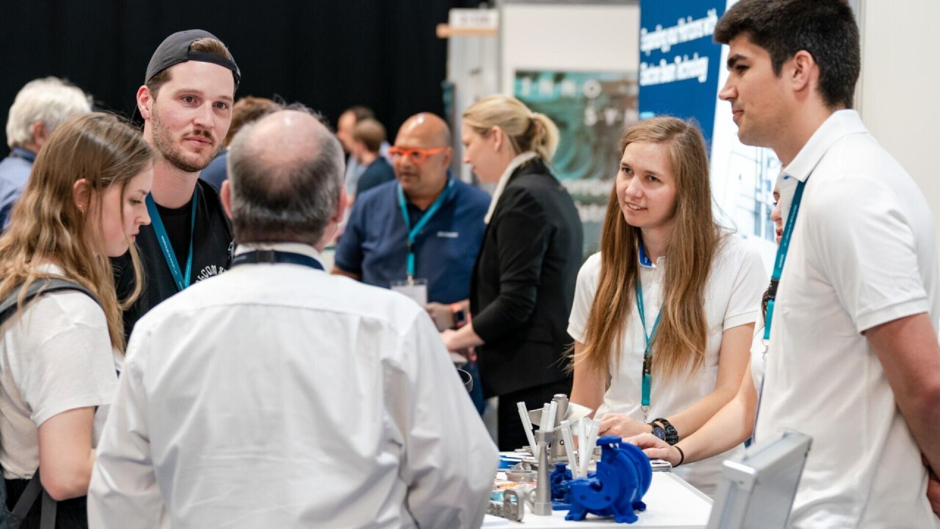 A group of people stand talking at a conference booth. Two women and one man in white shirts with blue lanyards are behind the table, engaging with visitors. Technical equipment and a display are visible on the table.