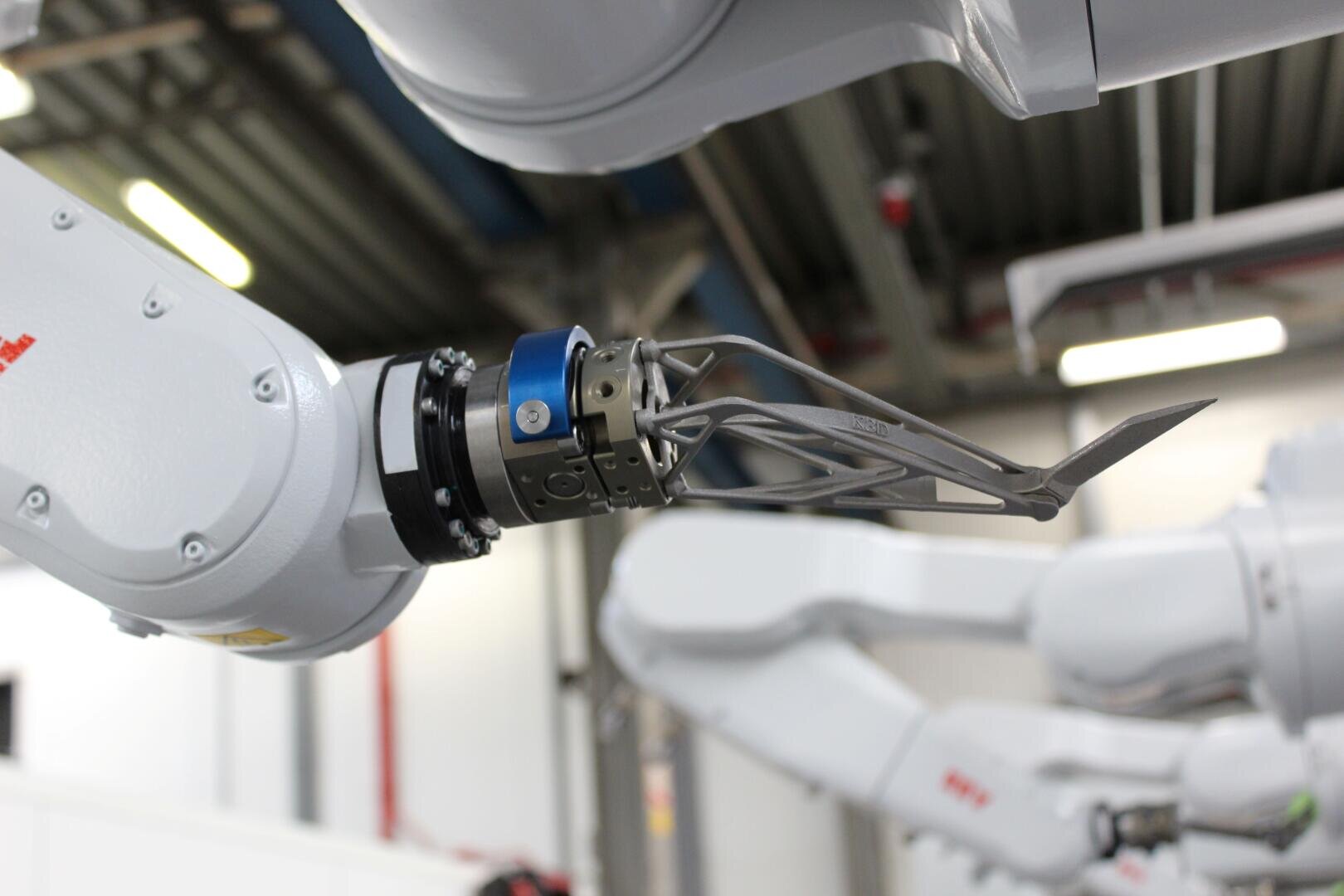 Eine Nahaufnahme eines Roboterarms mit einem Greiferaufsatz aus Metall in einer industriellen Umgebung, im Hintergrund ist ein weiterer Roboterarm zu sehen.