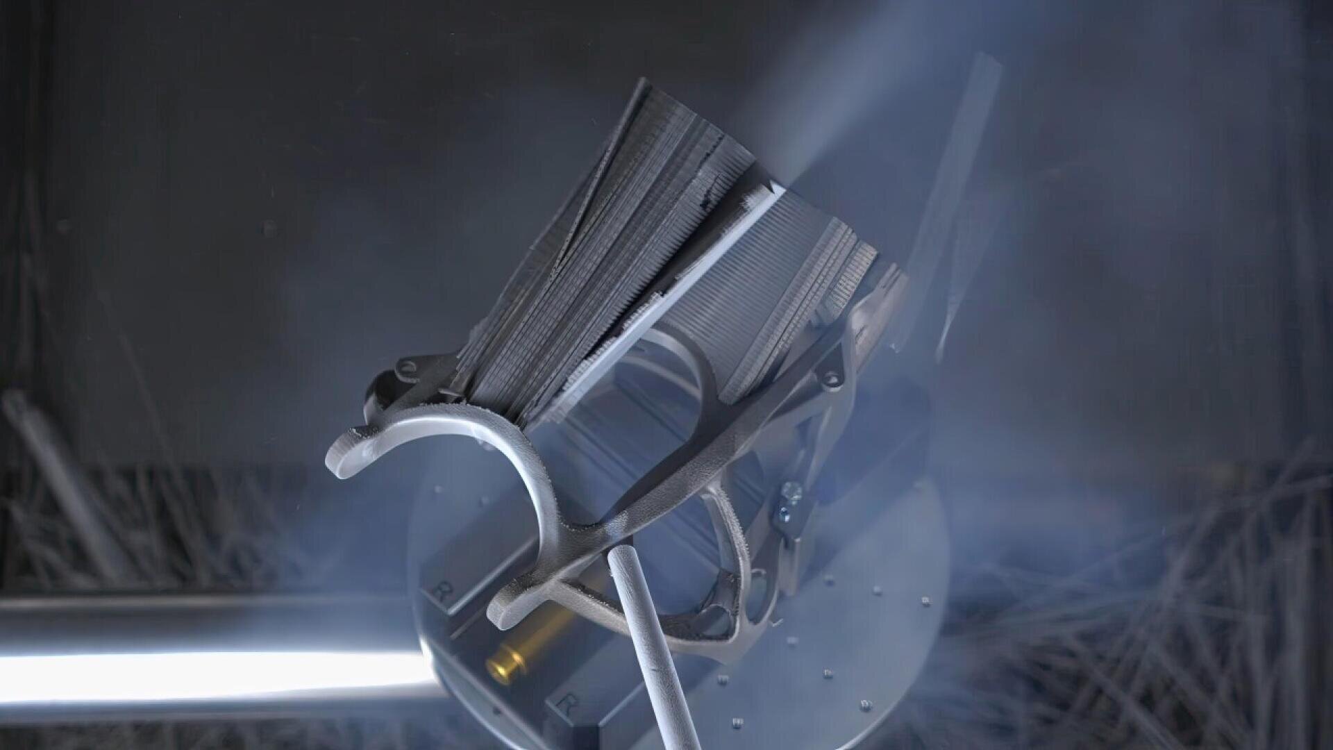 A metallic object is being held by a robotic arm, surrounded by mist or smoke, under focused beams of light in an industrial setting. The object appears to be a partially assembled or 3D-printed component.