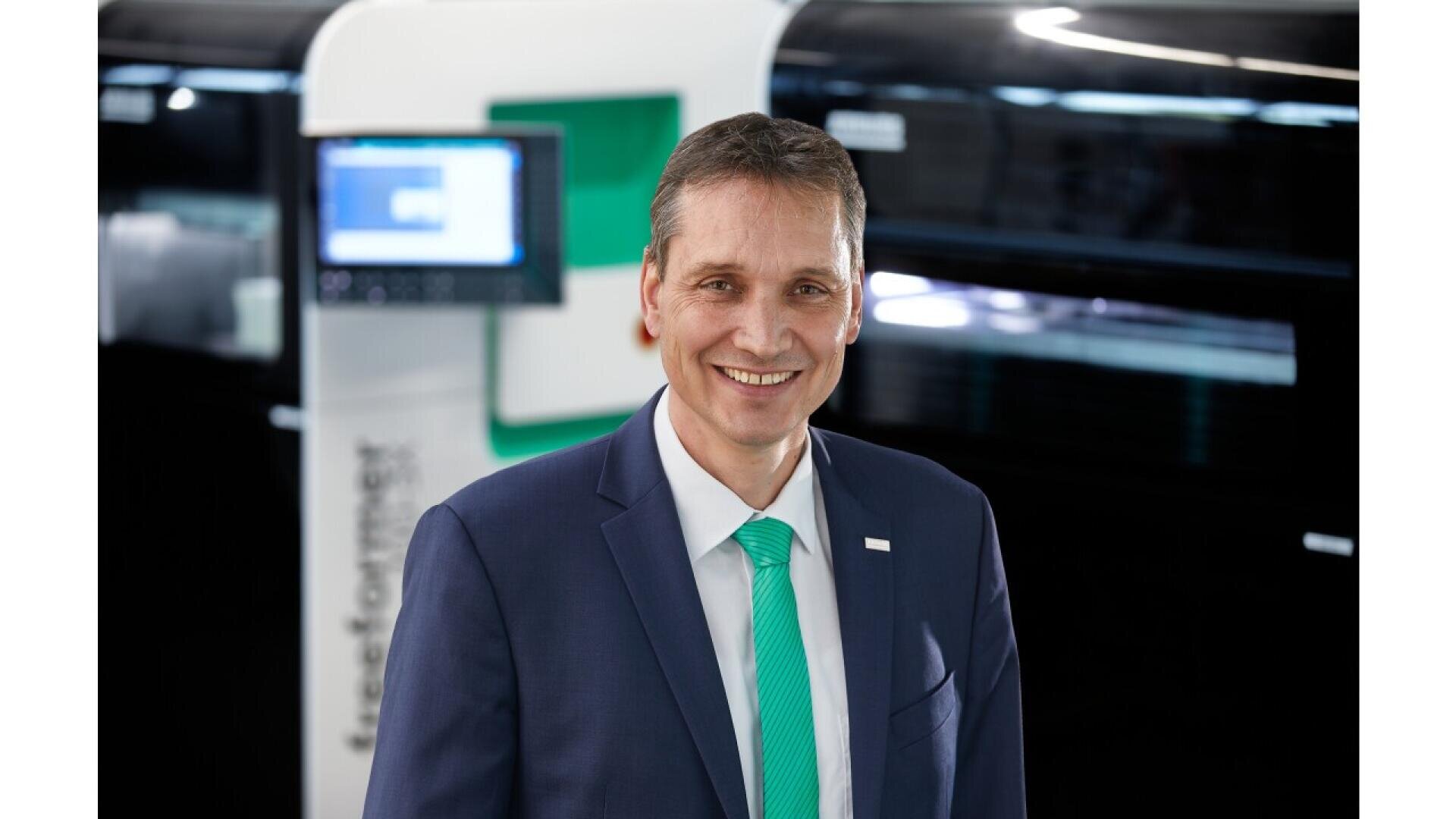 A man in a dark blue suit, white shirt, and teal tie smiles at the camera. He stands indoors in front of a modern, high-tech machine with a screen and blurred background.