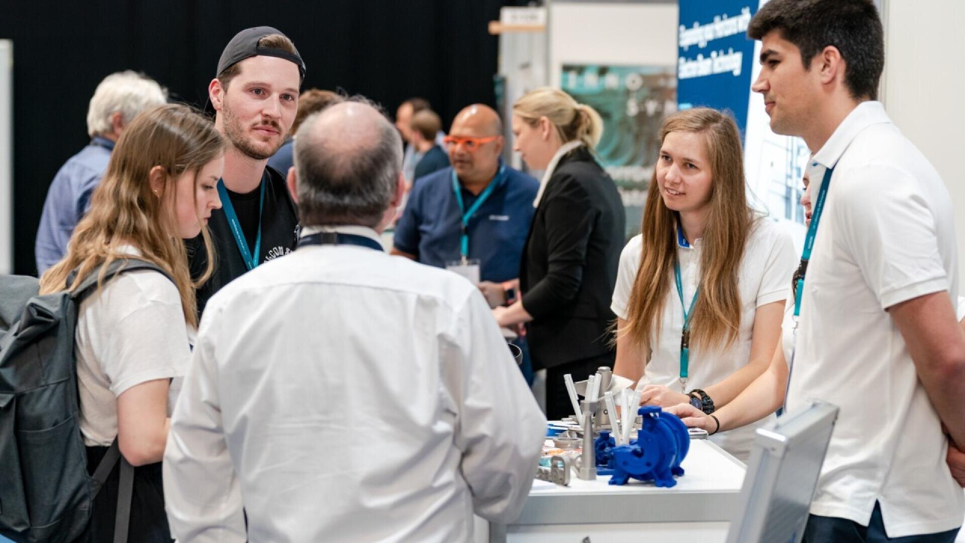 A group of people stand and talk at a trade show booth, with two women and a man behind a counter displaying items, while three attendees listen and engage in conversation. Other people are visible in the background.
