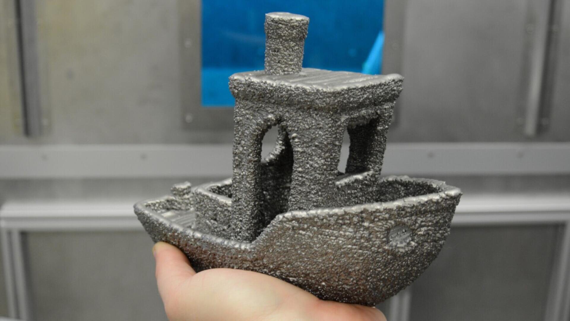A hand holds a small, 3D-printed boat model with a rough, metallic, textured surface. The background is blurred, showing a metallic wall and a blue window or screen.
