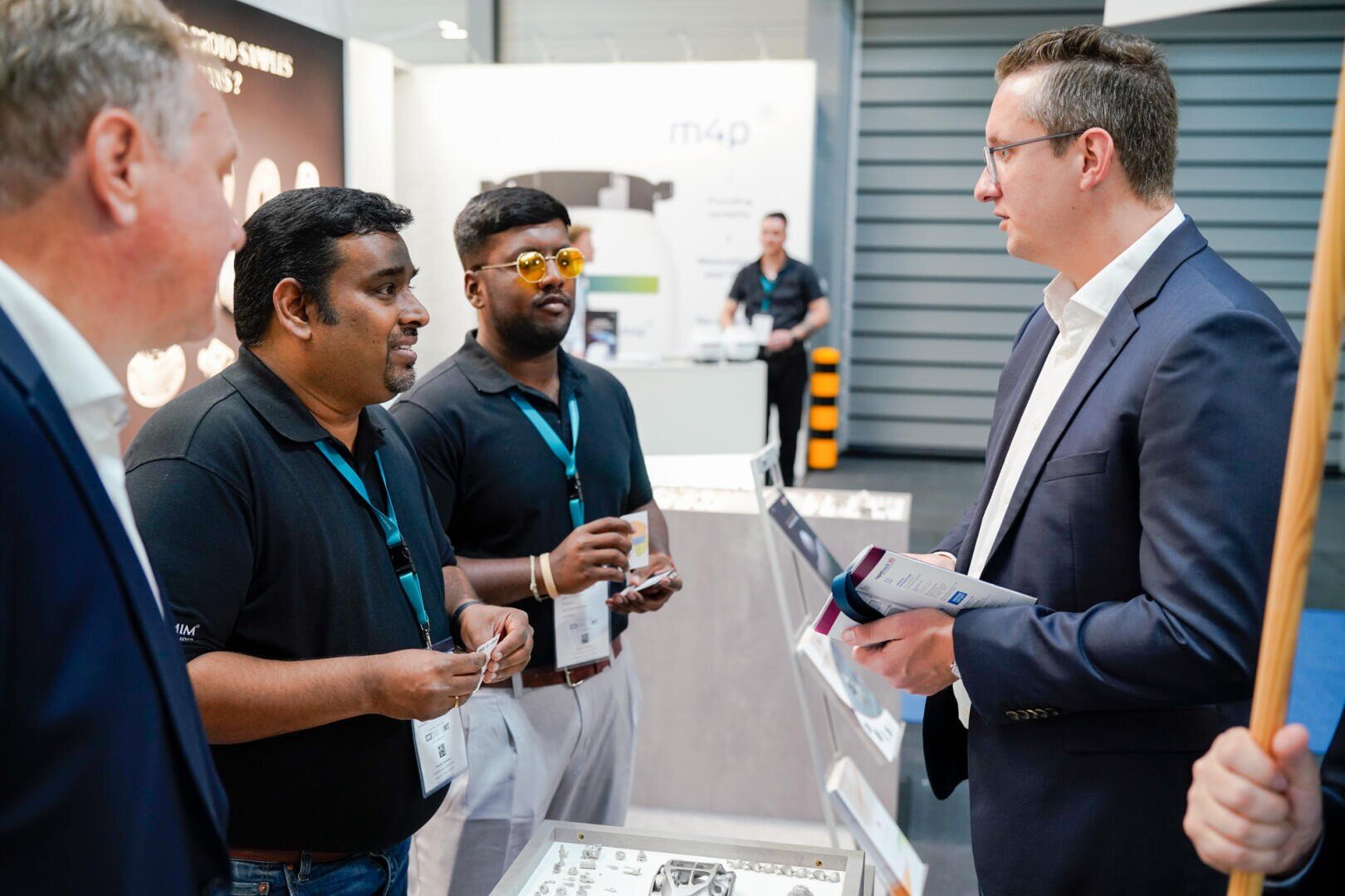 Vier Männer stehen auf einer Messe oder einer Geschäftsveranstaltung zusammen und unterhalten sich. Zwei Männer tragen schwarze Hemden und Schlüsselbänder, die anderen Anzüge. Um sie herum sind Stände und Informationsmaterialien zu sehen.