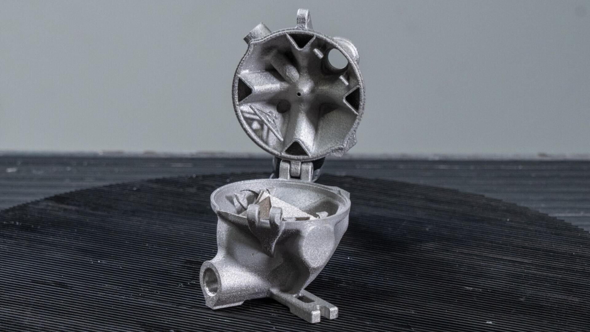 A small, metallic 3D-printed object resembling a toilet with an open lid sits on a ridged, dark surface against a plain gray background.