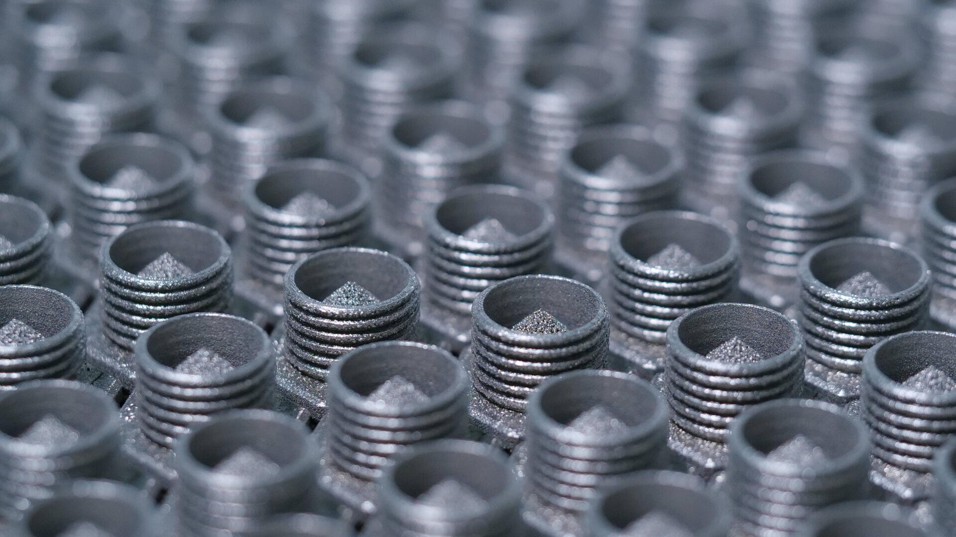 Reihen metallischer Gewindezylinder mit strukturierten Oberseiten, angeordnet in einem Gittermuster. Die Objekte haben eine glänzende, silberne Oberfläche und scheinen Maschinenteile oder Industriekomponenten zu sein.