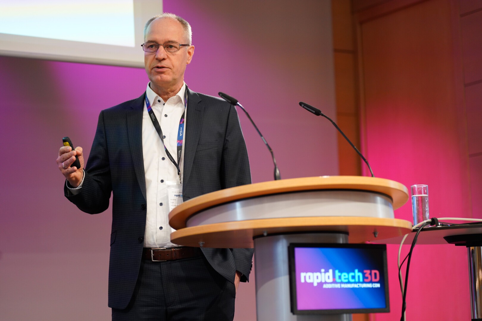 A man in a suit speaks at a podium with a microphone at the “rapid tech 3D” event. The background is lit with purple and pink lighting, and a glass of water sits on the podium.