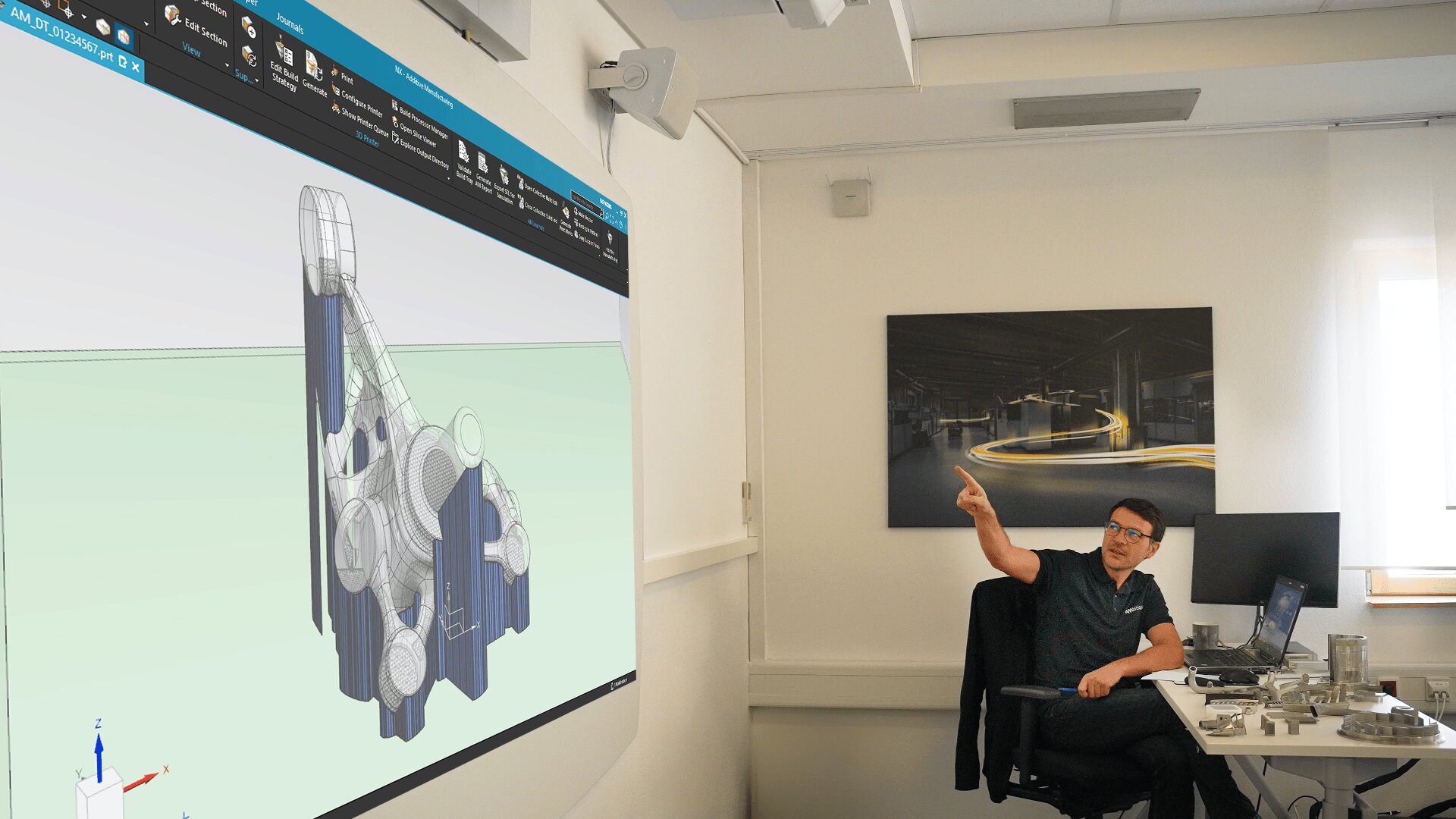 Eine Person sitzt an einem Schreibtisch in einem Büro und zeigt auf einen großen Bildschirm, auf dem ein 3D-Modell eines komplexen geometrischen Objekts angezeigt wird. Im Raum gibt es Computerausrüstung und ein Fenster.