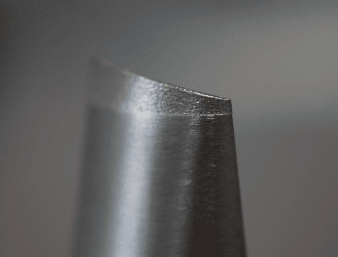 Eine Nahaufnahme einer scharfen, metallischen Schneidkante, möglicherweise eines Bohrers oder einer Klinge, mit einer polierten, abgewinkelten Spitze vor einem unscharfen grauen Hintergrund.