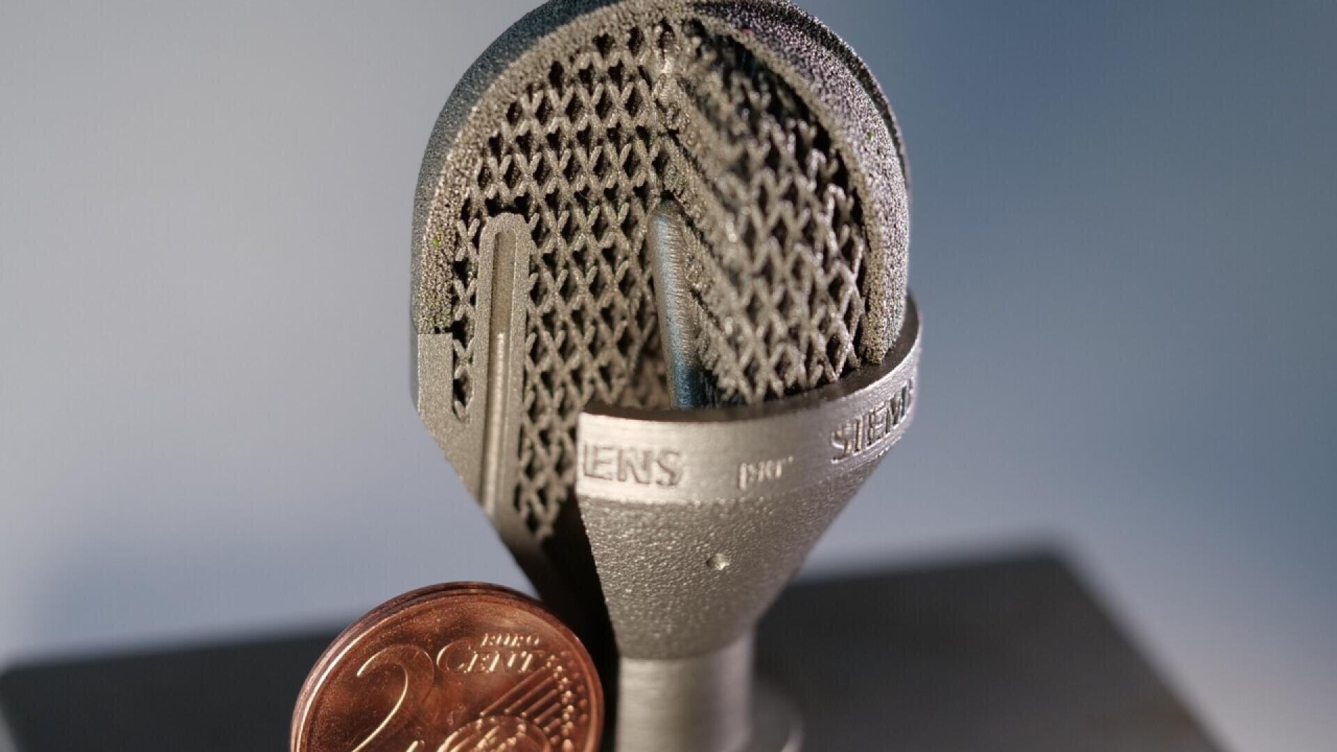 A close-up of a metallic 3D-printed object with a lattice structure, marked with SIEMENS, next to a 2-cent euro coin for scale, set against a blurred background.