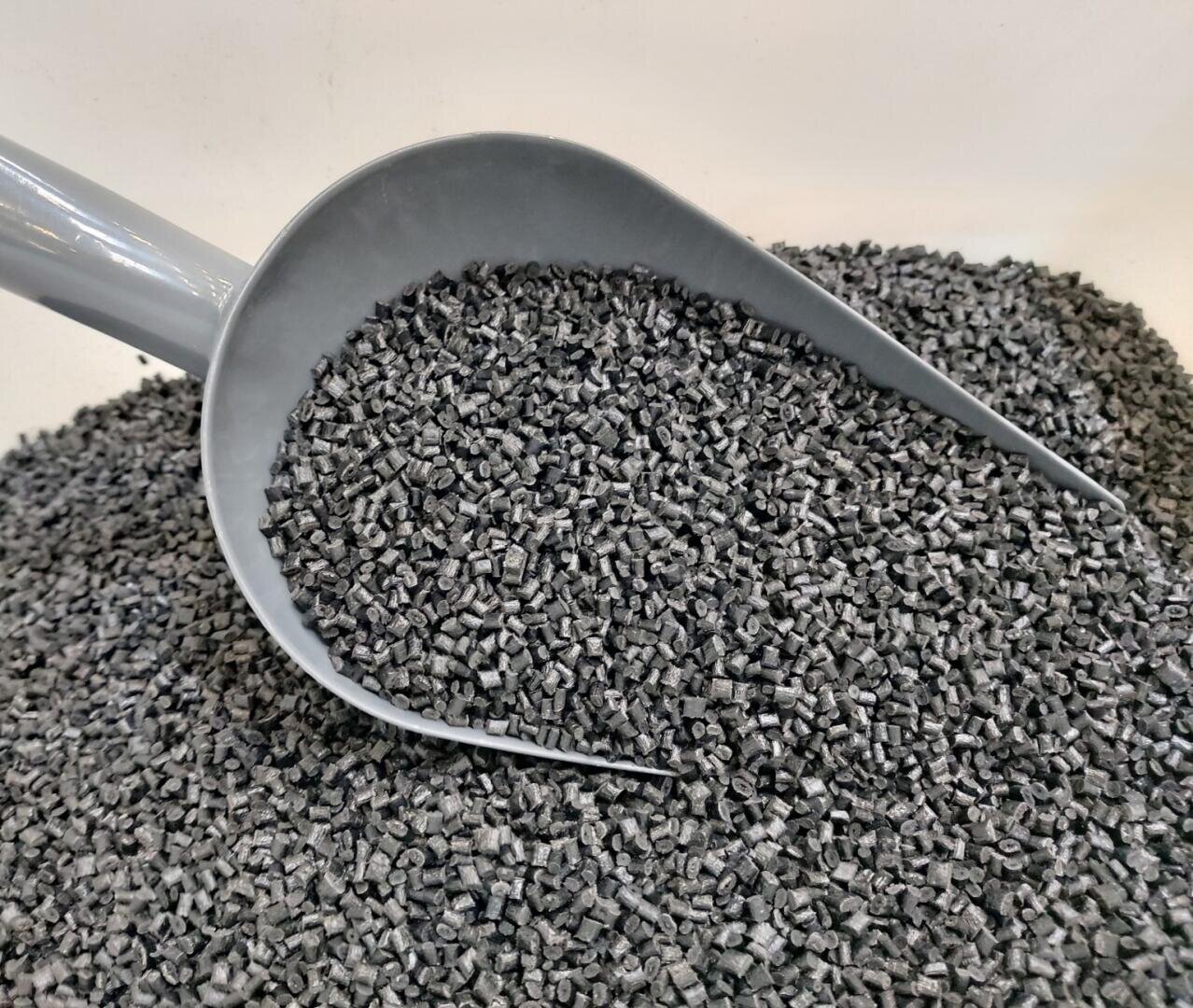 Eine graue Schaufel ist mit einem großen Haufen kleiner, schwarzer Kunststoffpellets gefüllt, die auf einer flachen Oberfläche verteilt sind.