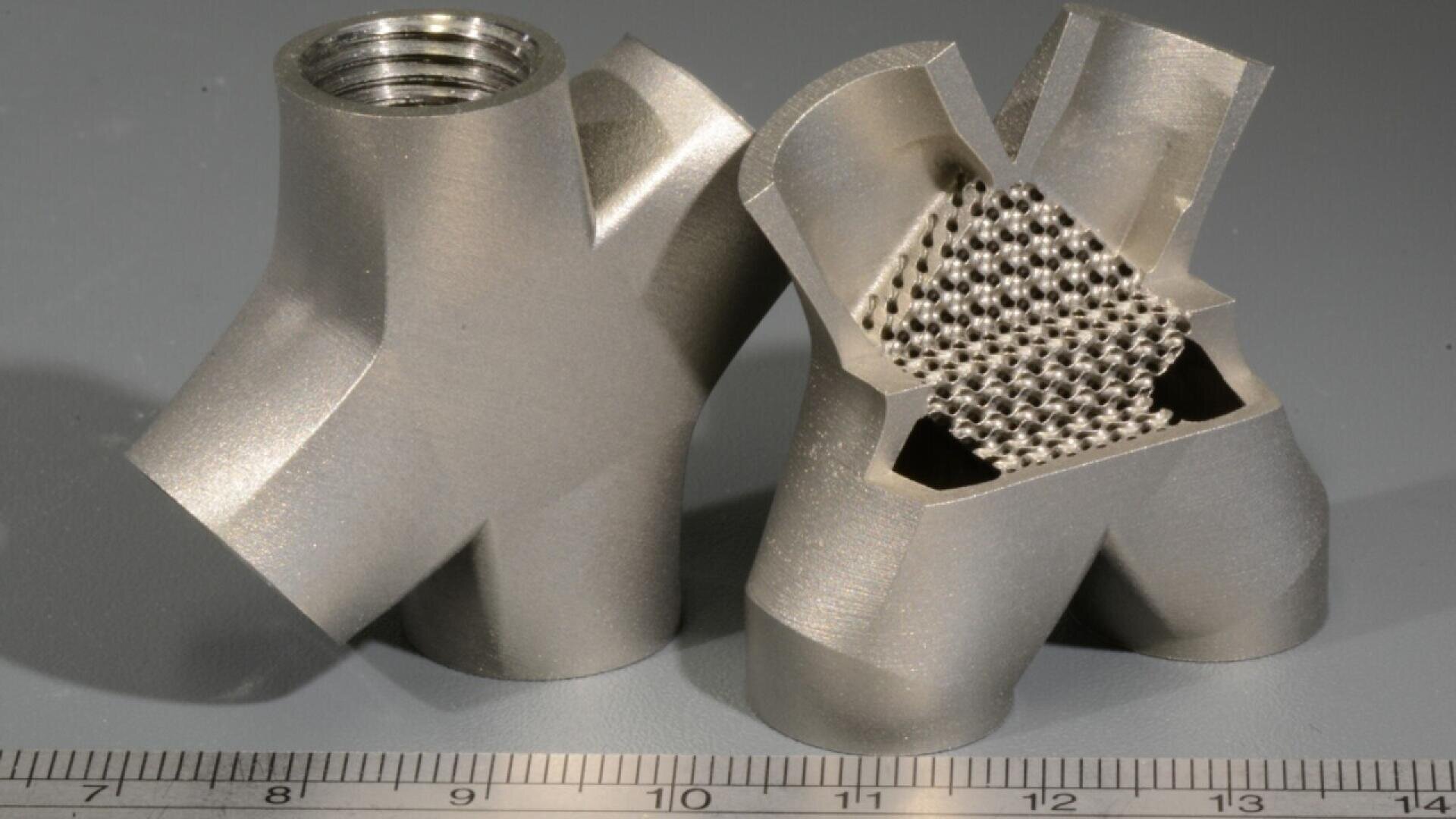Two metal 3D-printed pipe connectors are shown side by side; one is whole and the other is cut open, revealing a lattice structure inside. A ruler lies in front for scale.