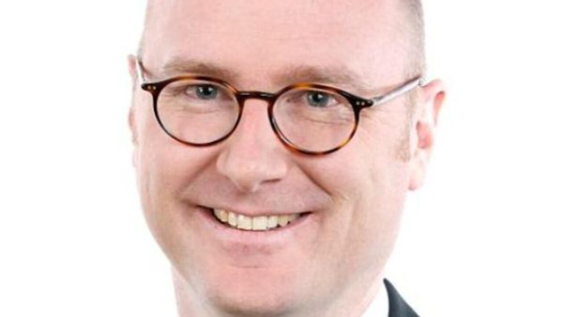 A smiling man with short light hair, wearing round eyeglasses and a suit, is pictured against a plain white background.