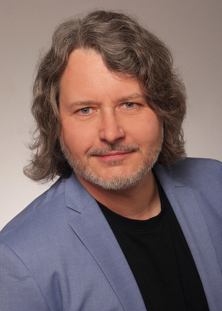 Ein Mann mit welligem grauem Haar und kurzem Bart, der einen hellblauen Blazer über einem schwarzen Hemd trägt, lächelt leicht, während er in die Kamera blickt. Der Hintergrund ist schlicht und hell.