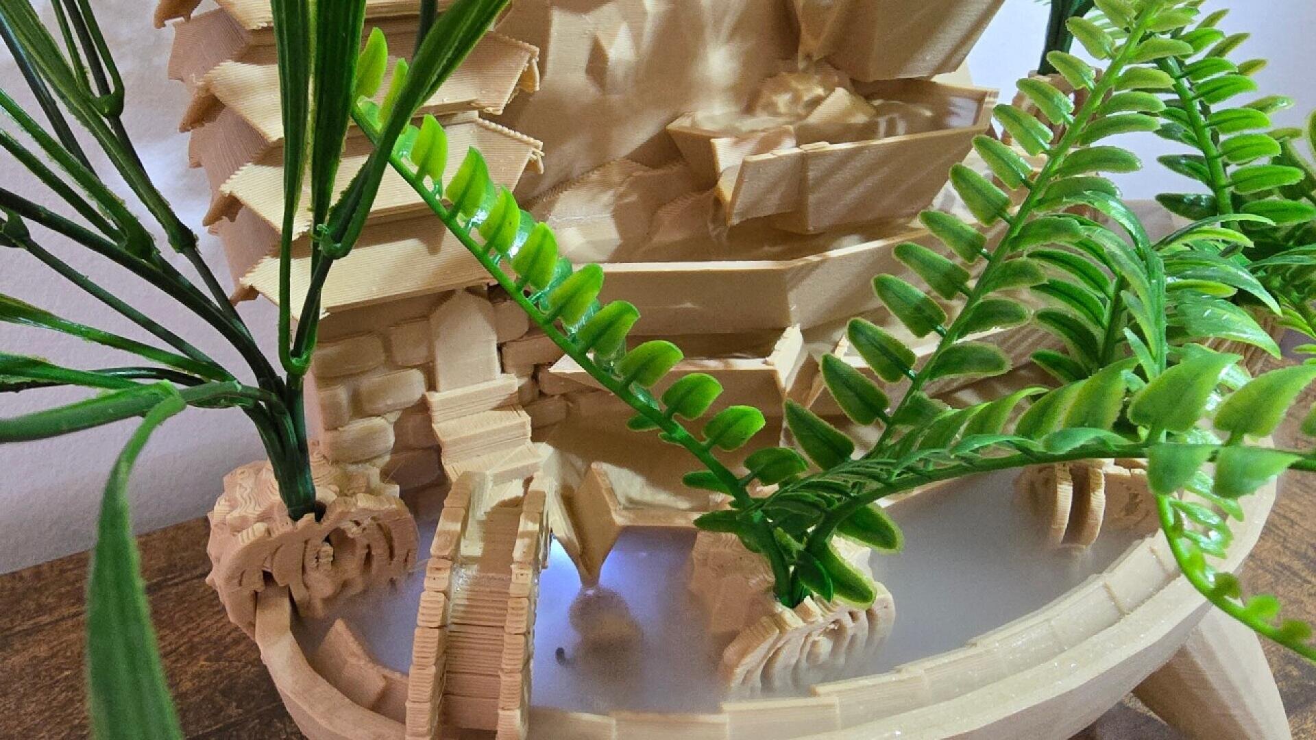 A close-up of a small wooden or 3D-printed model of a watermill with a waterwheel, surrounded by green artificial plants, placed on a wooden surface.