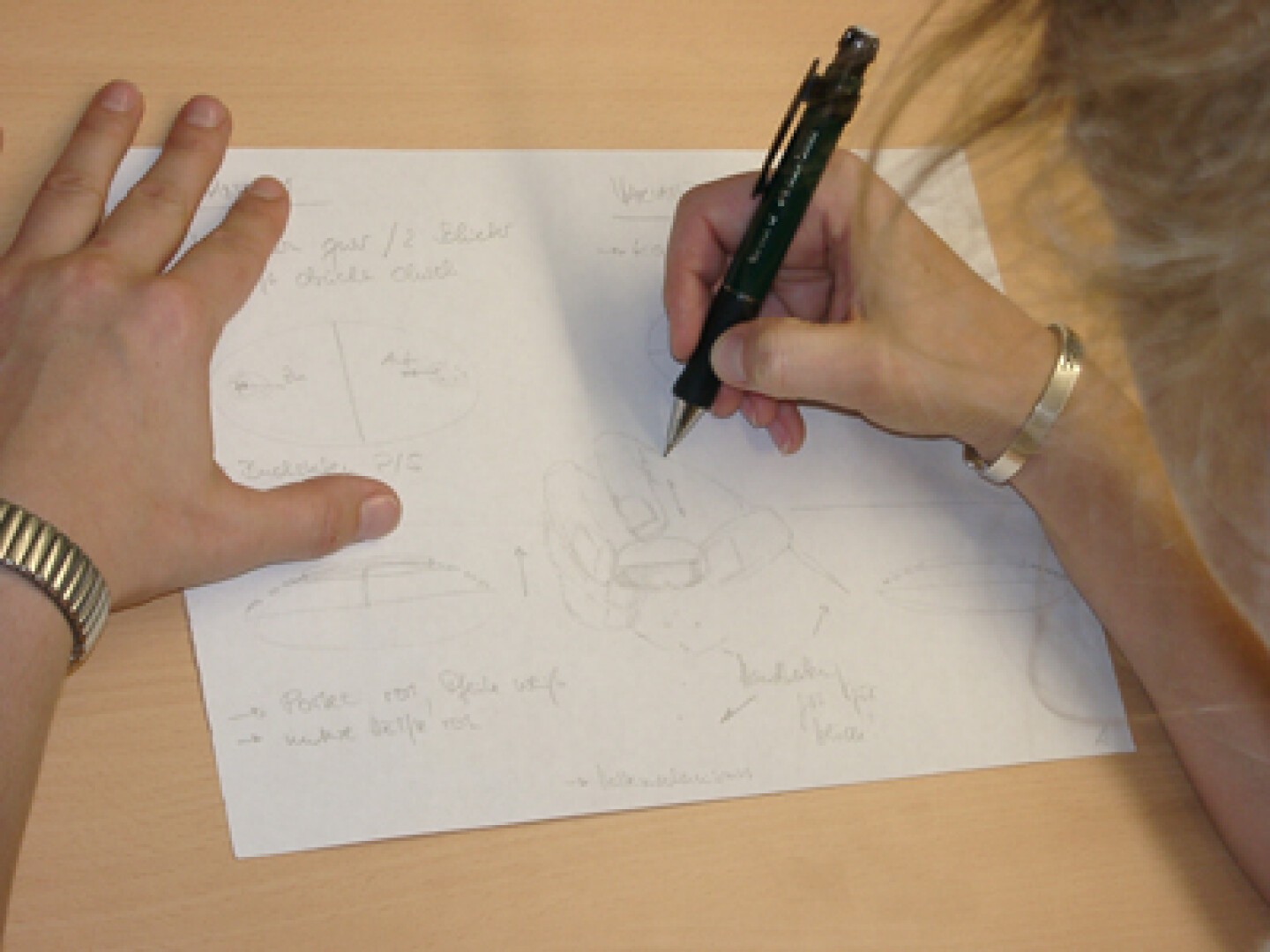Eine Person mit einer Uhr skizziert mit einem Stift Designkonzepte auf einem Blatt Papier. Das Papier zeigt grobe Zeichnungen und handschriftliche Notizen und liegt auf einem hellen Holzschreibtisch.