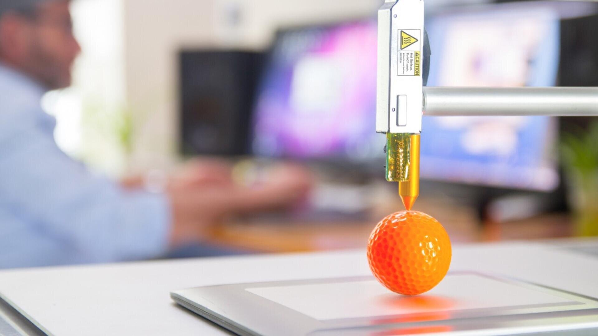 A close-up of a 3D printer nozzle printing an orange golf ball on a build plate, with a person using a computer in the blurred background.
