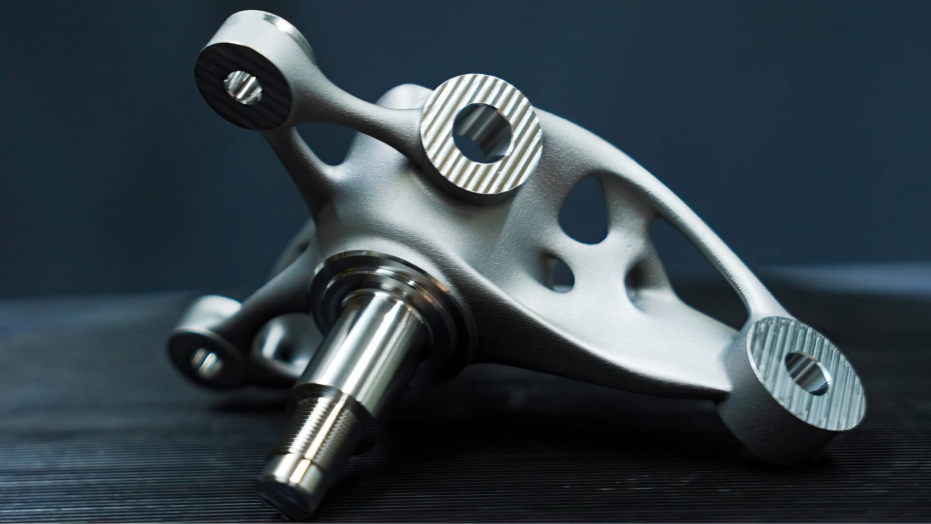 Eine Nahaufnahme eines metallischen, kompliziert gestalteten mechanischen Teils mit glatten und strukturierten Oberflächen, kreisförmigen Löchern und einer zentralen Welle, das auf einer dunklen Oberfläche vor einem dunklen Hintergrund ruht.