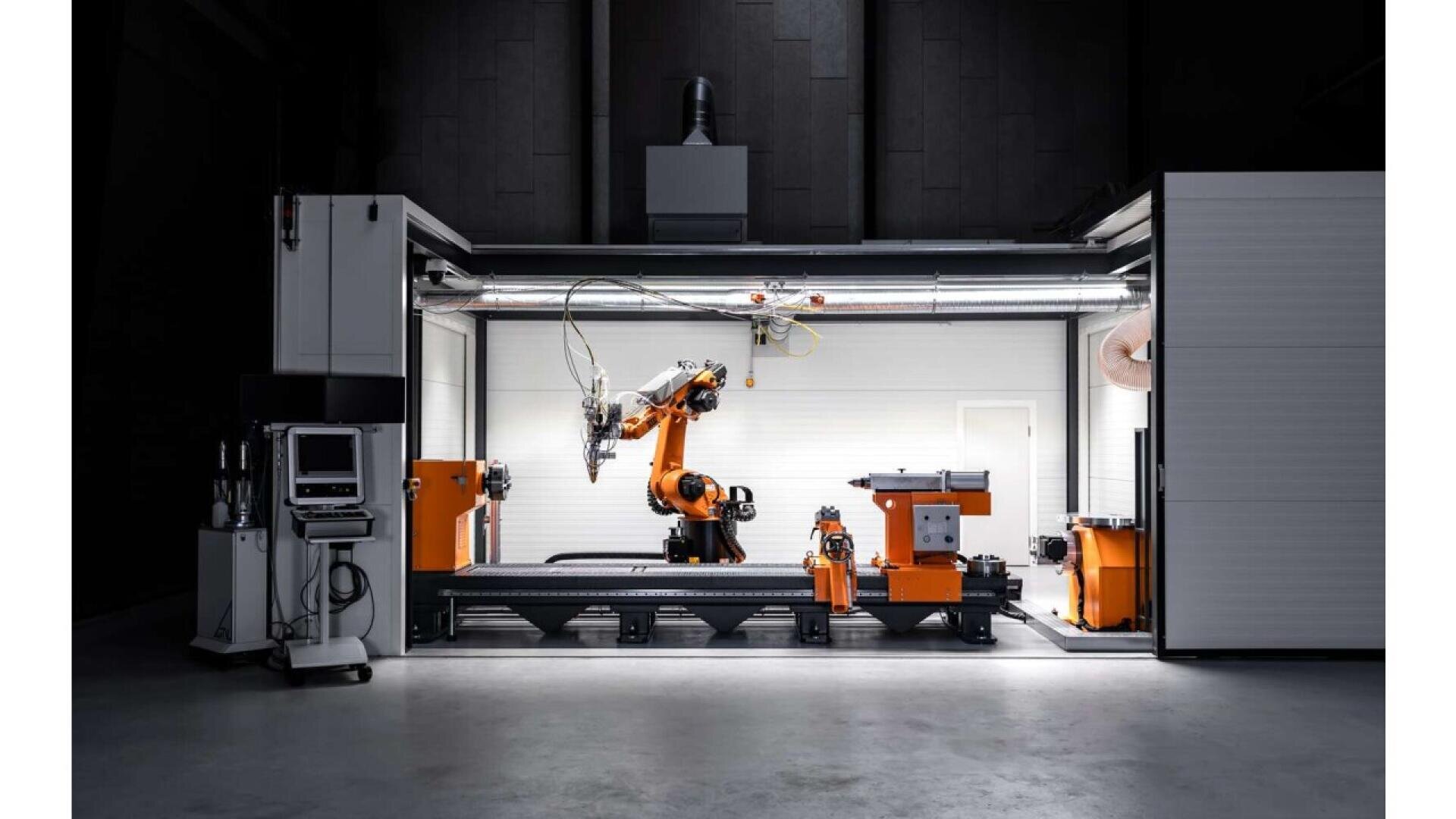 A modern industrial robotic arm in an enclosed workspace with orange and black machinery, surrounded by equipment and computers, illuminated by overhead lights in a high-tech manufacturing environment.