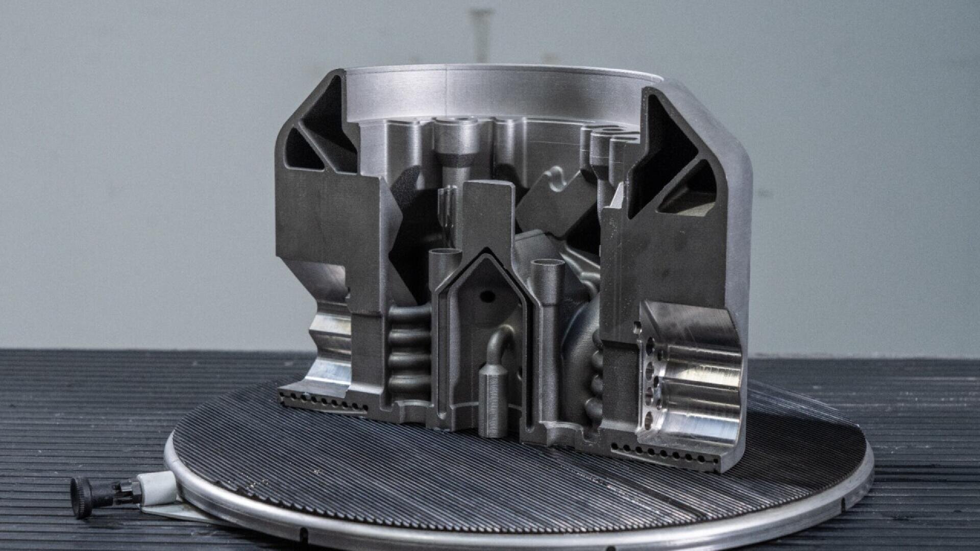 A metallic cylindrical component with a detailed internal structure is displayed on a round platform, partially cut to reveal intricate inner channels and geometric features.