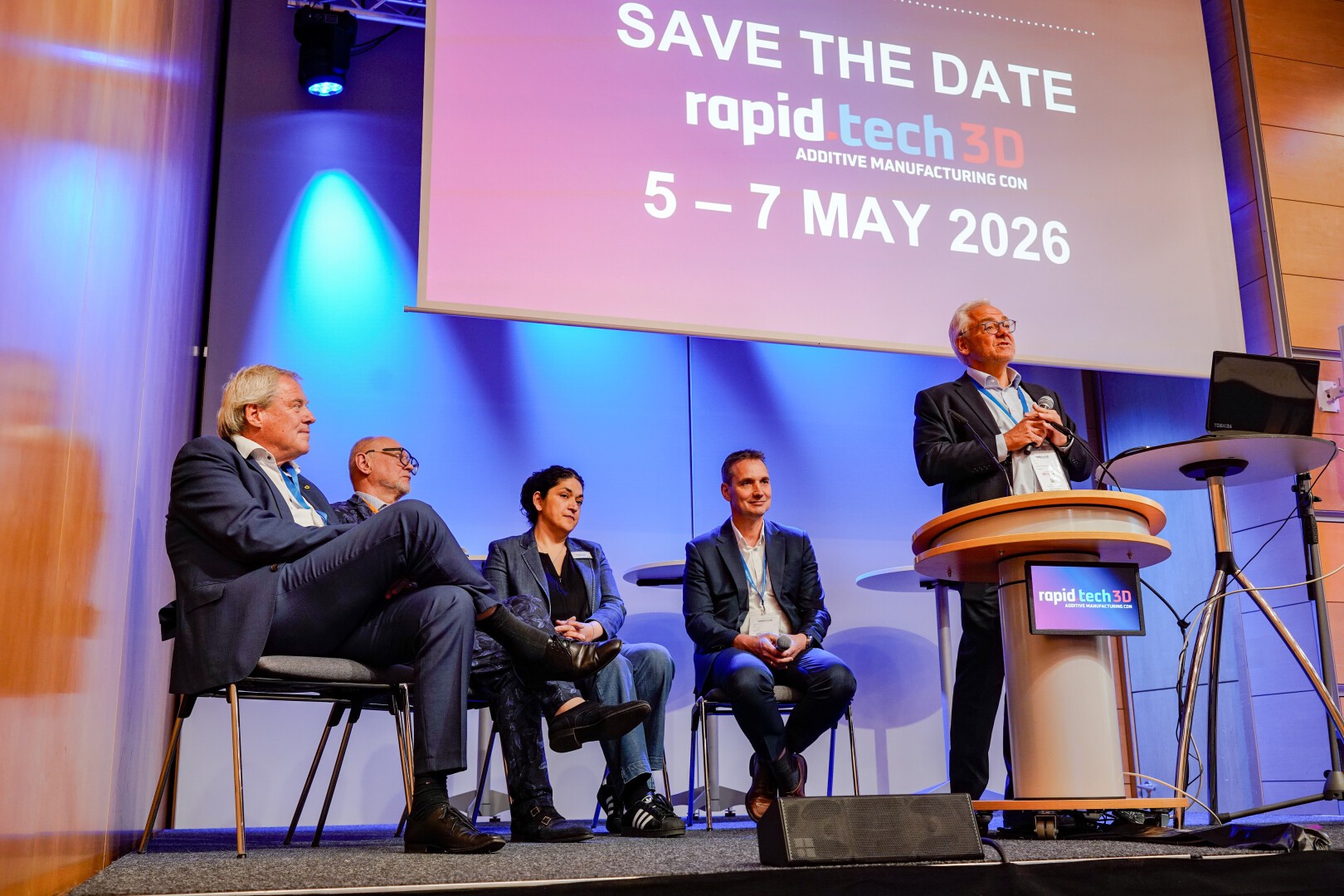 Fünf Personen in Businesskleidung sitzen und stehen auf einer Konferenzbühne. Über ihnen steht auf einem großen Schild: „Merken Sie sich den Termin für die Rapid Tech 3D ADDITIVE MANUFACTURING CON 5.–7. Mai 2026“. Ein Mann hält eine Rede auf einem Podium.