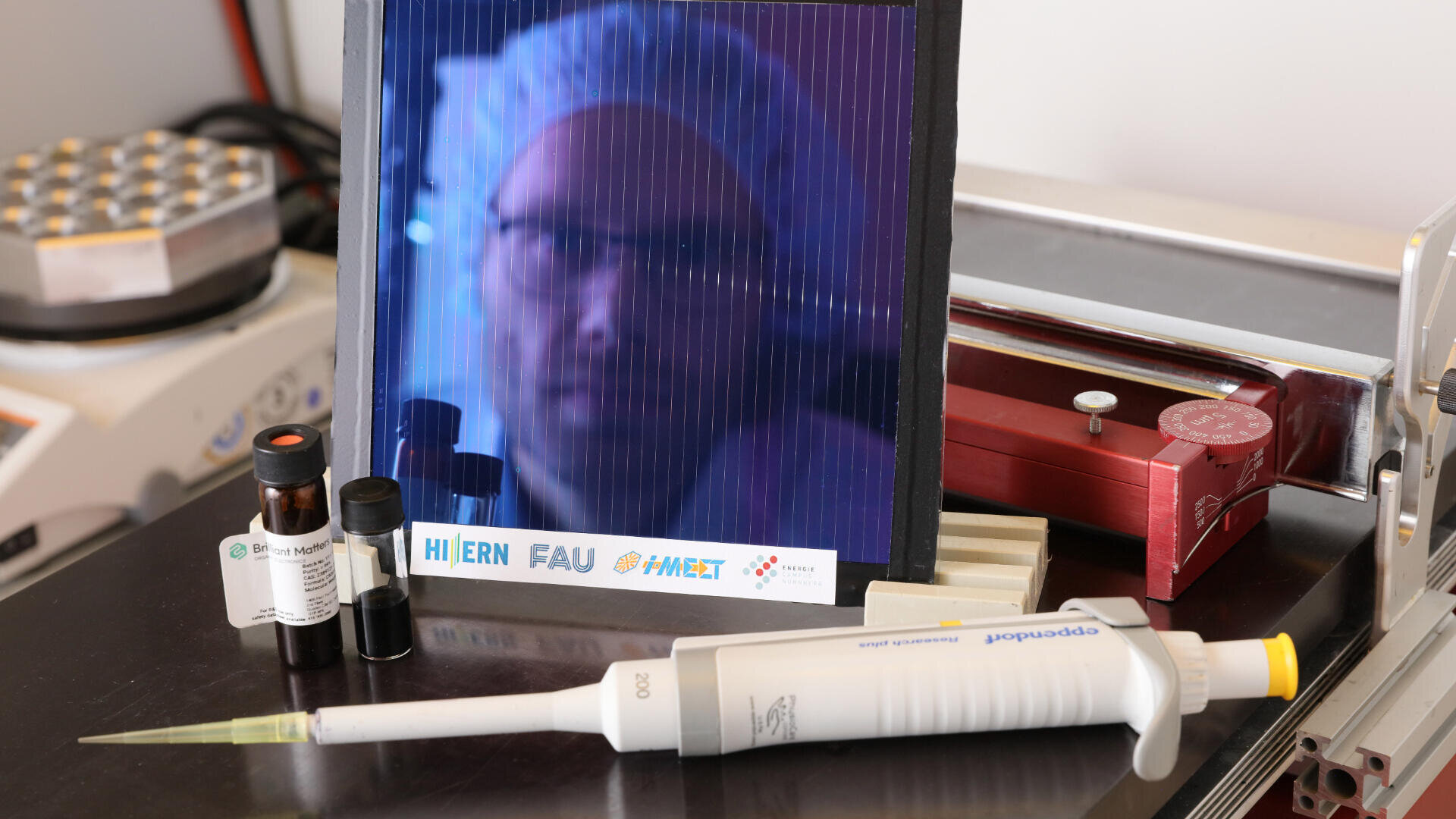 A close-up of a lab bench with a pipette, vials, and equipment. A reflective surface shows a blurred person in a lab coat and hairnet. Various science labels and instruments are visible.