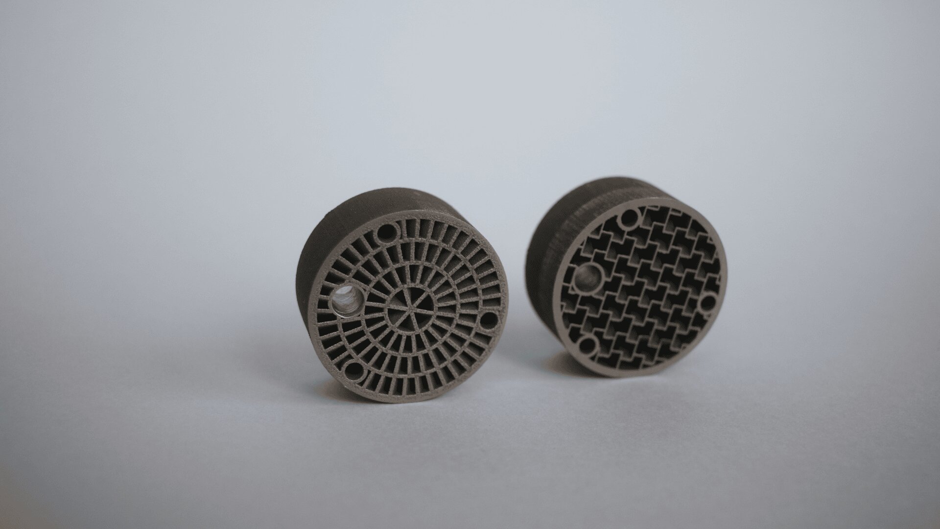 Zwei runde, graue 3D-gedruckte Objekte mit komplizierten Mustern auf der Vorderseite werden auf eine helle Fläche gelegt. Das linke Objekt hat ein radiales Muster, während das rechte Objekt ein gitterartiges Muster aufweist.