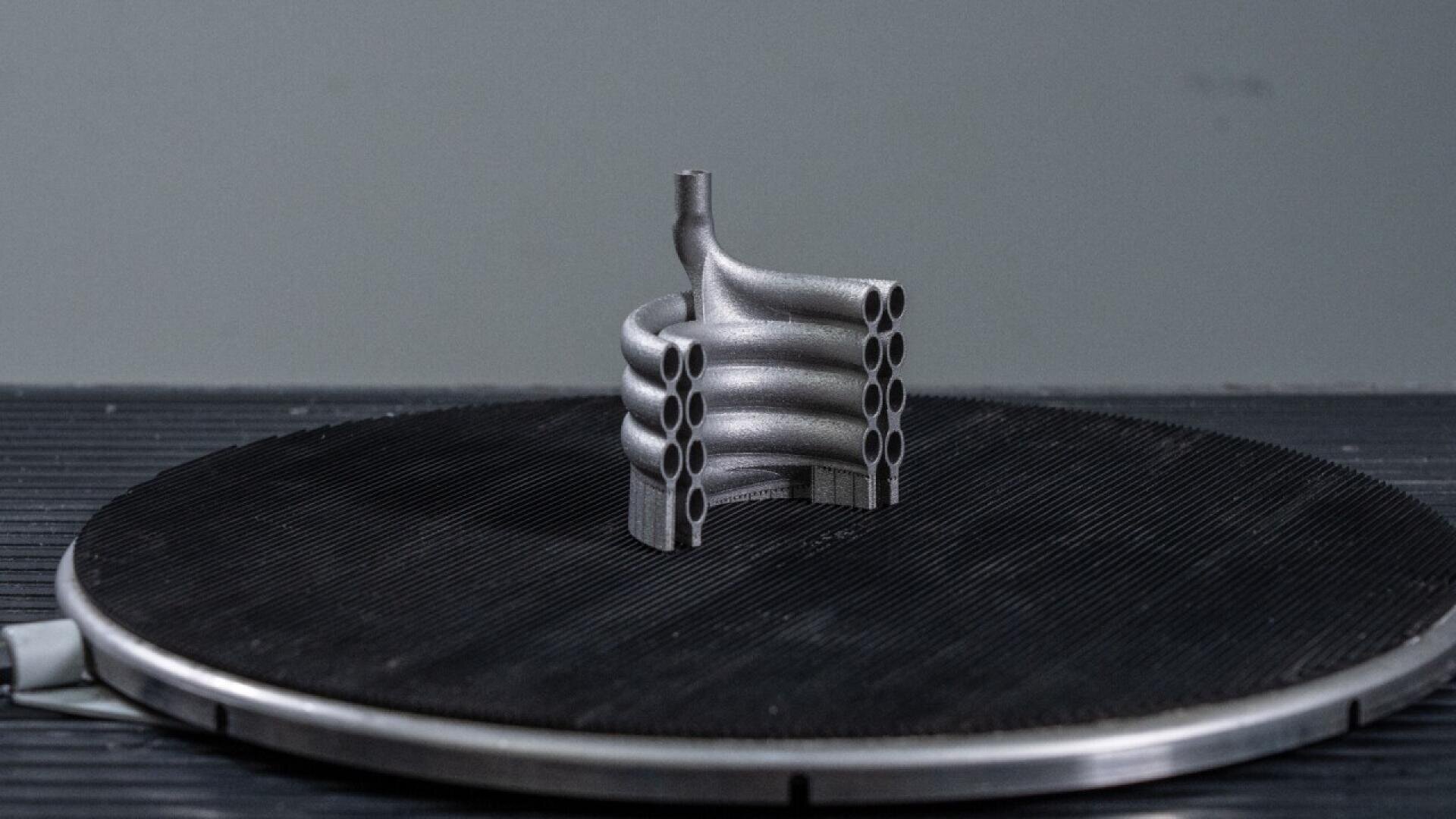 A metallic 3D-printed component with multiple curved, tubular structures is displayed upright on a circular, black textured platform against a plain gray background.
