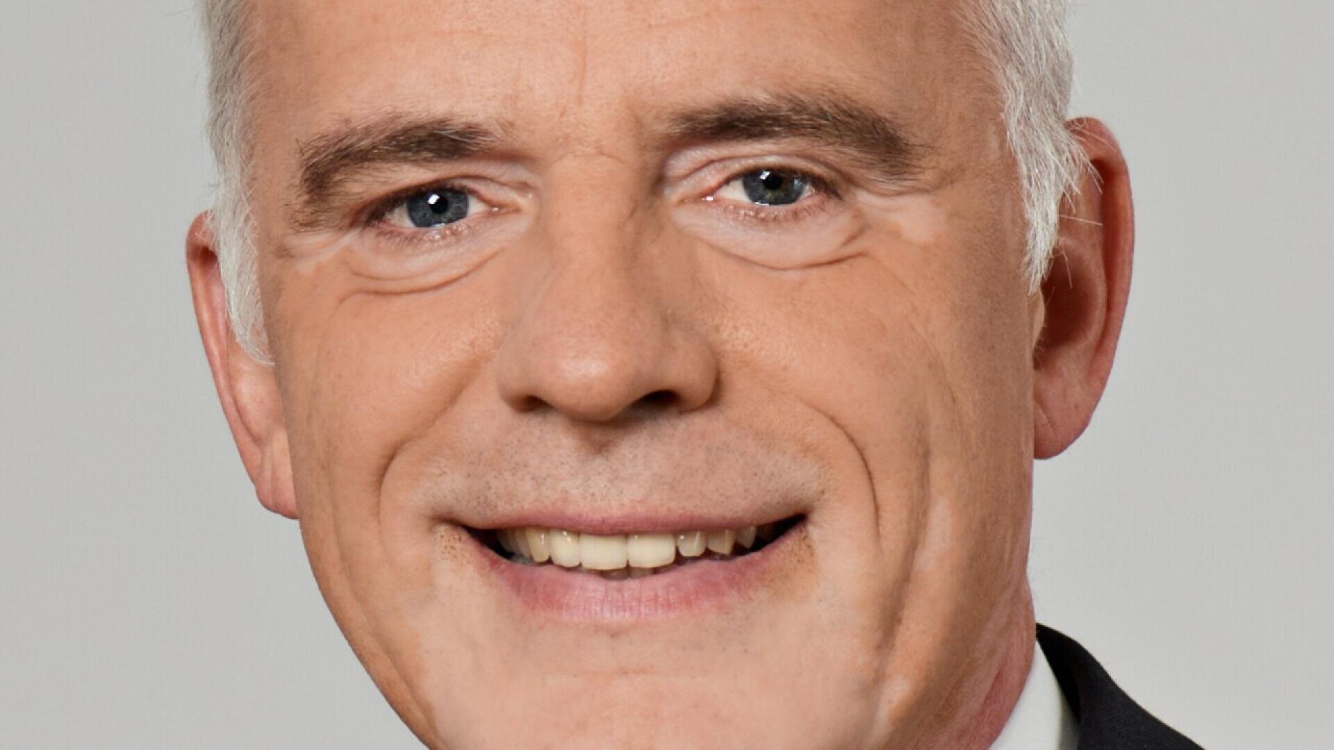Close-up of a middle-aged man with light skin, short gray hair, blue eyes, and a friendly smile, wearing a dark suit and white shirt against a plain light background.