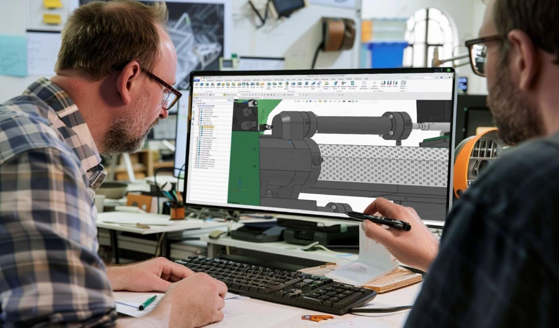 Zwei Personen arbeiten in einem modernen Büro gemeinsam an einem Schreibtisch. Sie blicken auf einen Computerbildschirm, auf dem eine 3D-Software zur mechanischen Konstruktion mit der technischen Zeichnung eines Maschinenteils angezeigt wird. Während der Diskussion gestikuliert einer mit einem Stift.