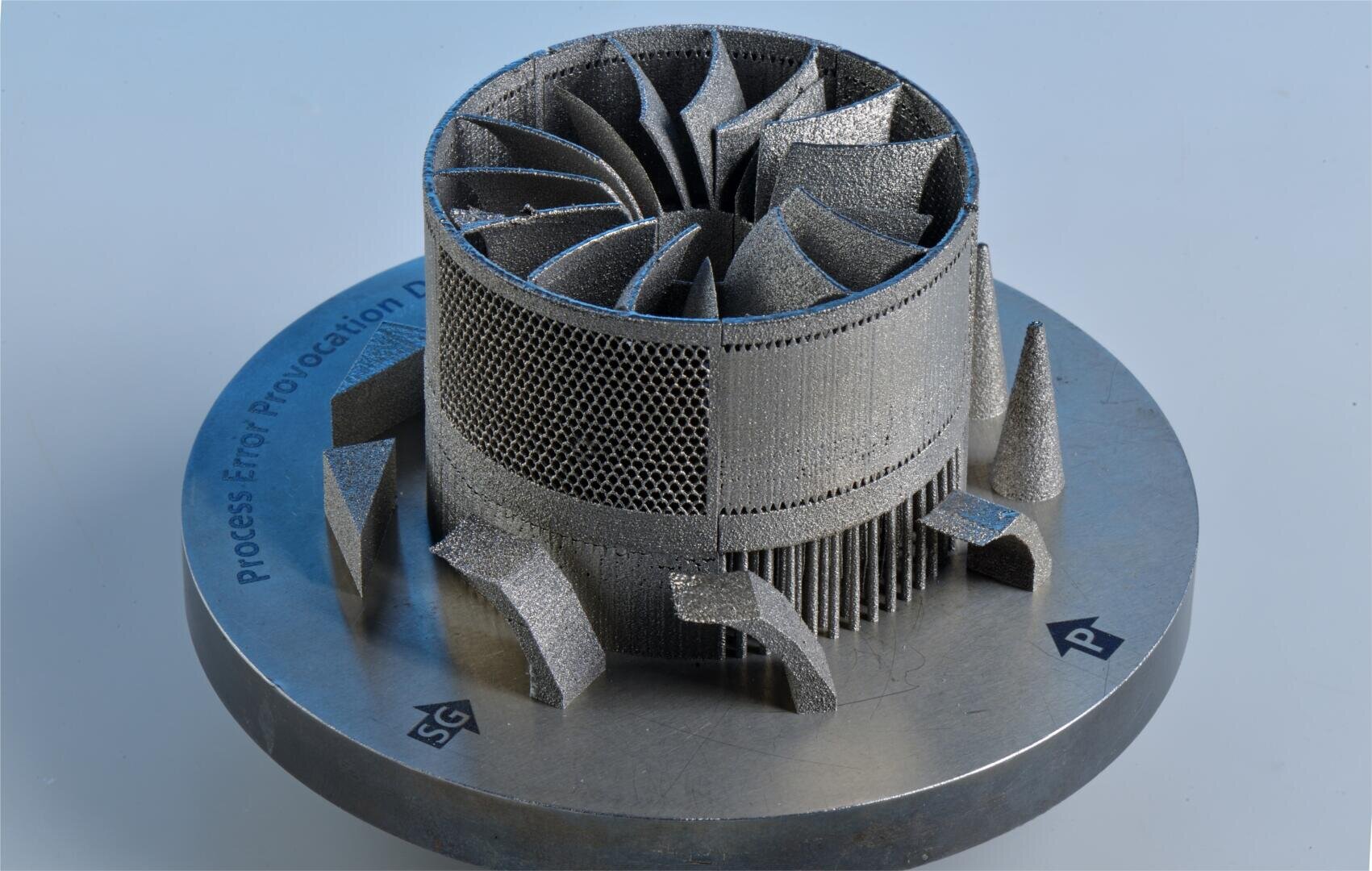 Ein metallisches, 3D-gedrucktes Turbinenbauteil mit verschlungenen Schaufeln und Gitterstrukturen sitzt auf einem runden Sockel, der mit Symbolen und Text beschriftet ist und auf einer hellen Fläche steht.