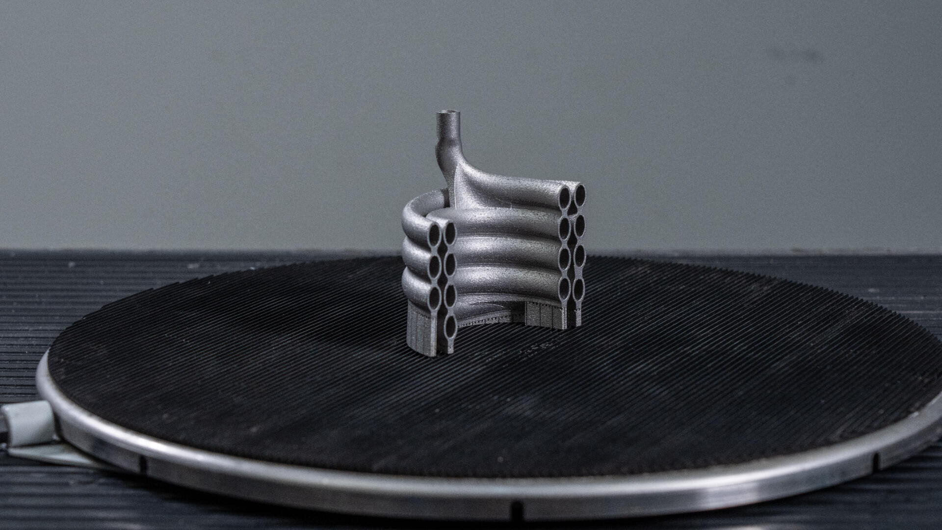 Eine metallische, 3D-gedruckte Komponente mit mehreren gebogenen, parallelen Rohren und einem zentralen vertikalen Rohr sitzt auf einer strukturierten, kreisförmigen schwarzen Plattform vor einem schlichten grauen Hintergrund.
