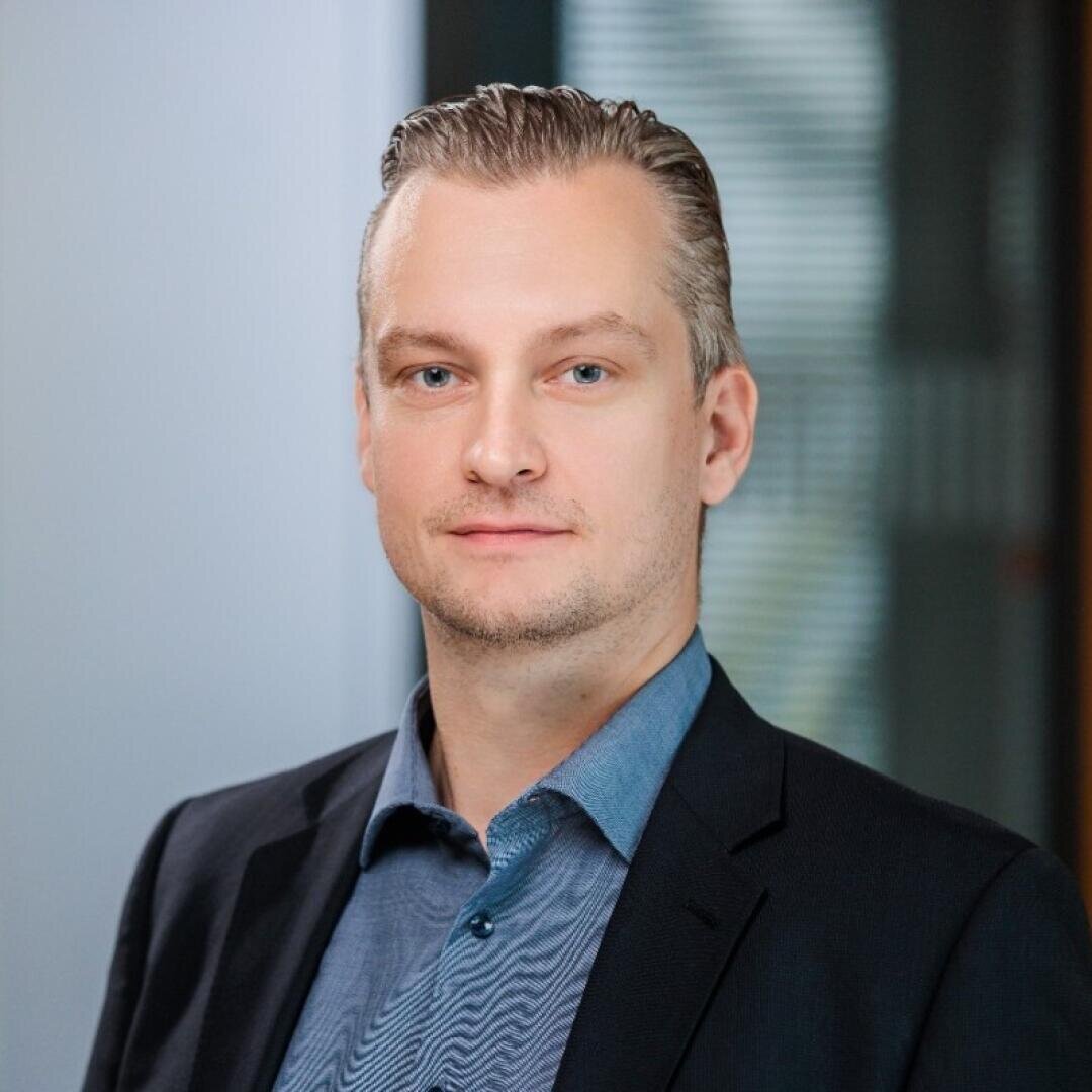Ein Mann mit kurzem, zurückgekämmtem hellem Haar und heller Haut trägt ein blaues Hemd und einen dunklen Blazer und steht in einem Gebäude vor einem unscharfen Bürohintergrund.