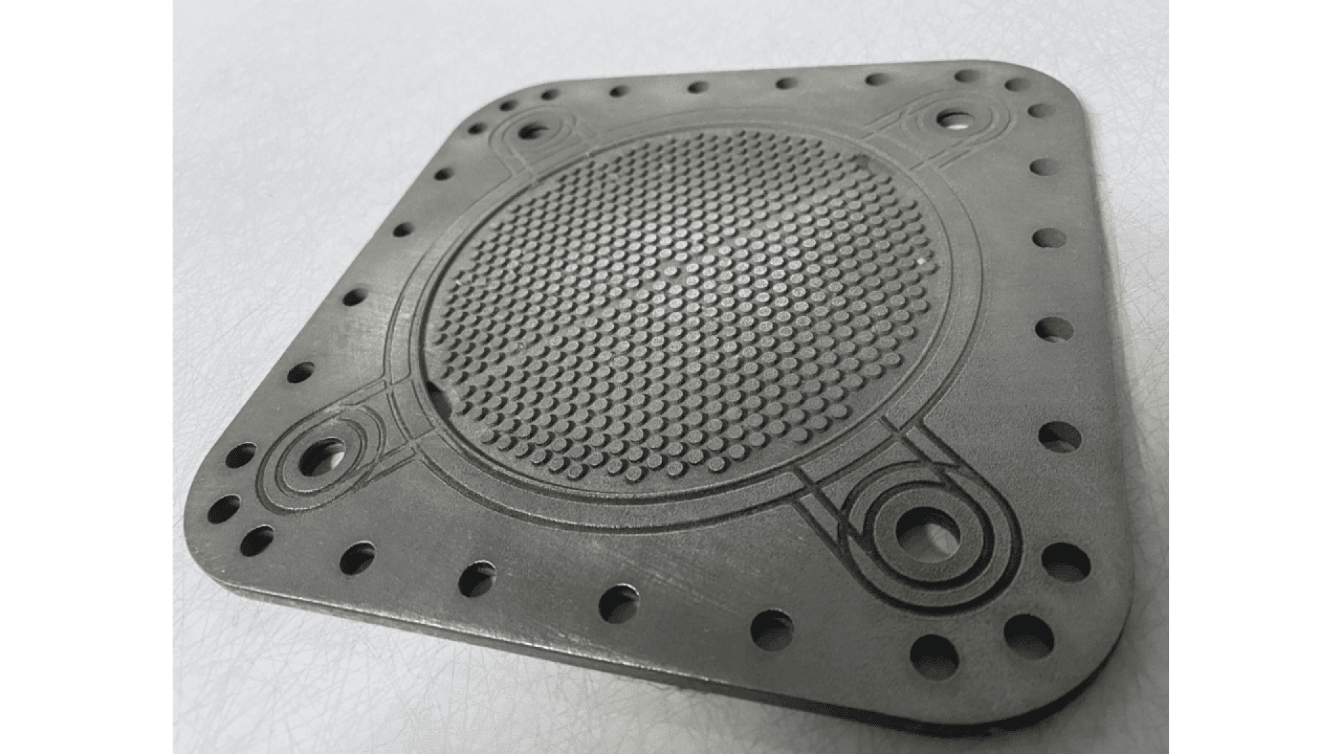 A close-up view of a square metal component with rounded corners, circular grooves, four large corner holes, and multiple smaller holes along the edges. The center area features a raised, textured grid pattern.