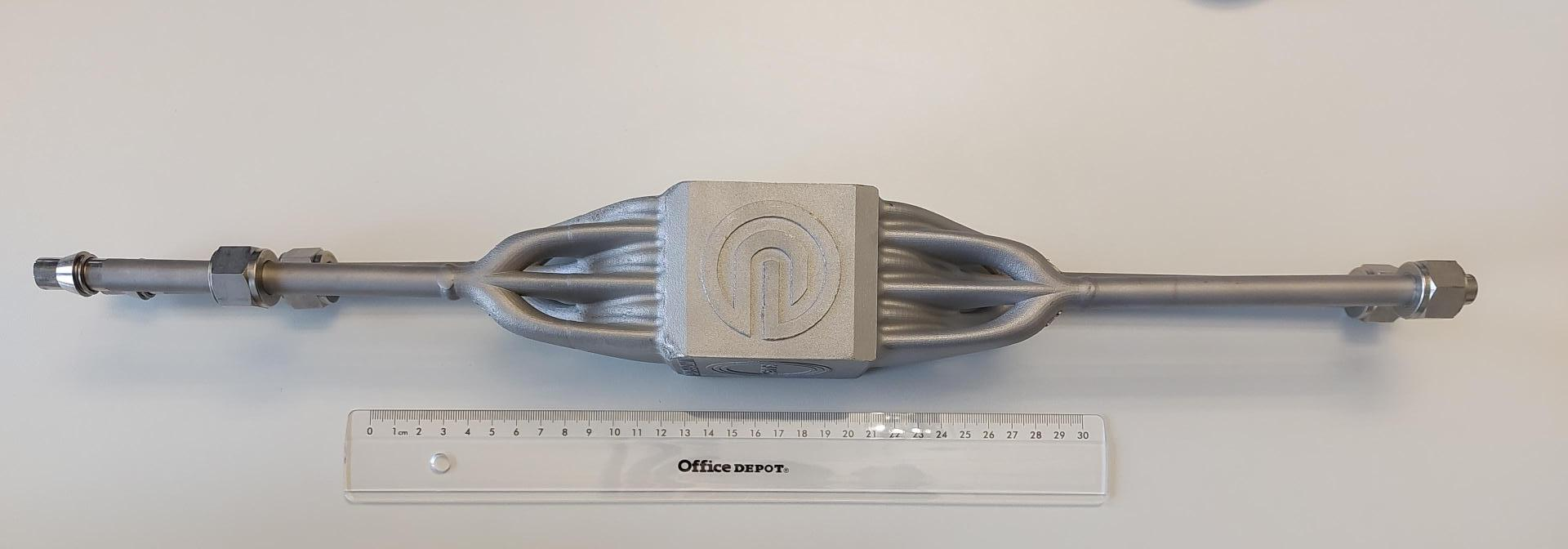 A metallic, tubular mechanical component with a square center and hexagonal ends is placed above a 30 cm clear plastic ruler on a white surface. The component features a stylized logo on the center block.