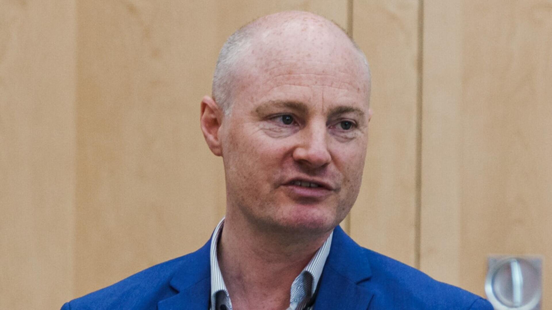 A man with fair skin, short thinning hair, wearing a blue suit jacket and striped shirt, stands in front of a light wood-paneled wall, looking slightly to the side.