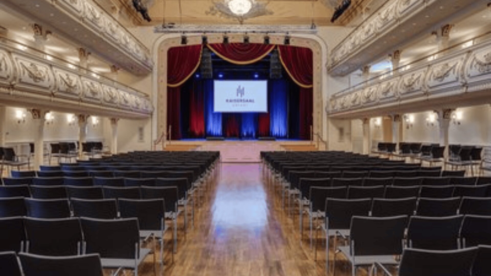 Ein großer, eleganter Saal mit schwarzen Stuhlreihen vor einer Bühne. Die Bühne hat rote Vorhänge, einen blau beleuchteten Hintergrund und eine Leinwand, auf der das Wort Kaisersaal steht. Verschnörkelte Balkone säumen die Seiten.