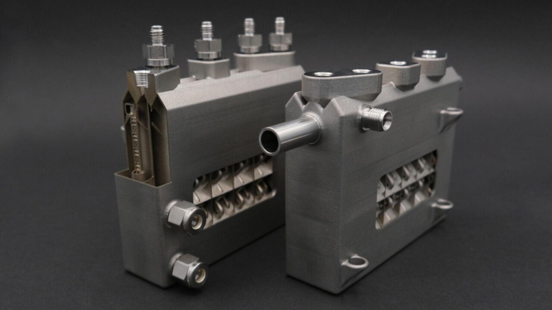 Two metallic, rectangular mechanical components with visible bolts and threaded connectors are displayed on a dark background. One component has an extended cylindrical pipe and both show intricate internal structures.
