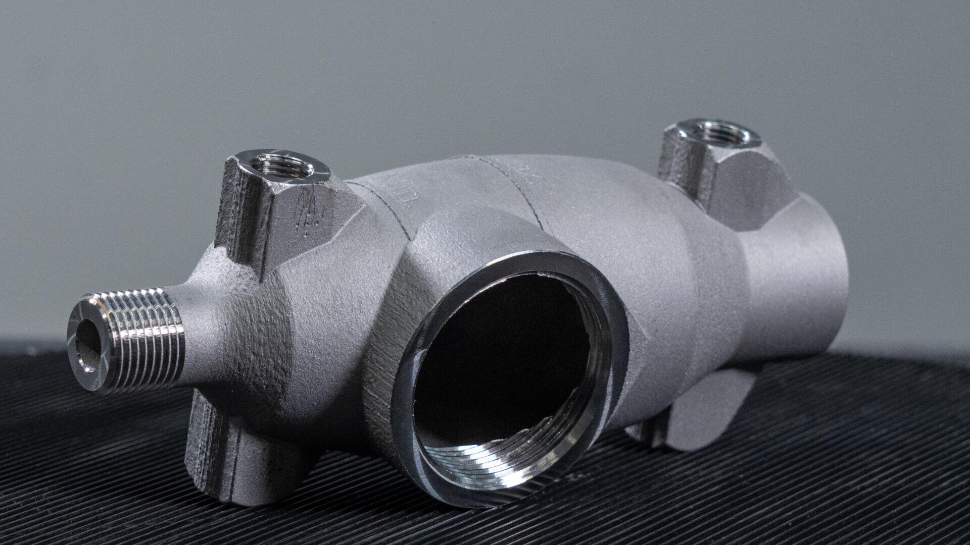 Eine Nahaufnahme einer metallischen Industrierohrverbindung mit Gewindeöffnungen an mehreren Enden, platziert auf einer dunklen strukturierten Oberfläche mit einem schlichten grauen Hintergrund.