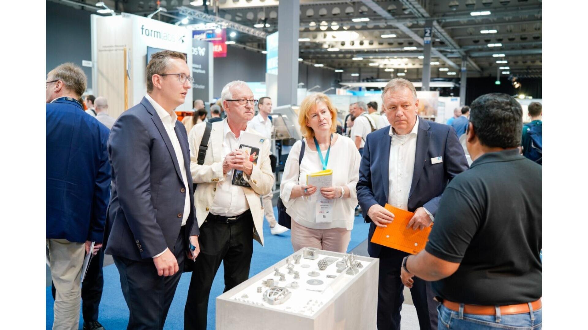 A group of professionals stands around a display table with metal parts at a busy trade show or convention, engaging in discussion. Exhibition booths and other attendees are visible in the background.