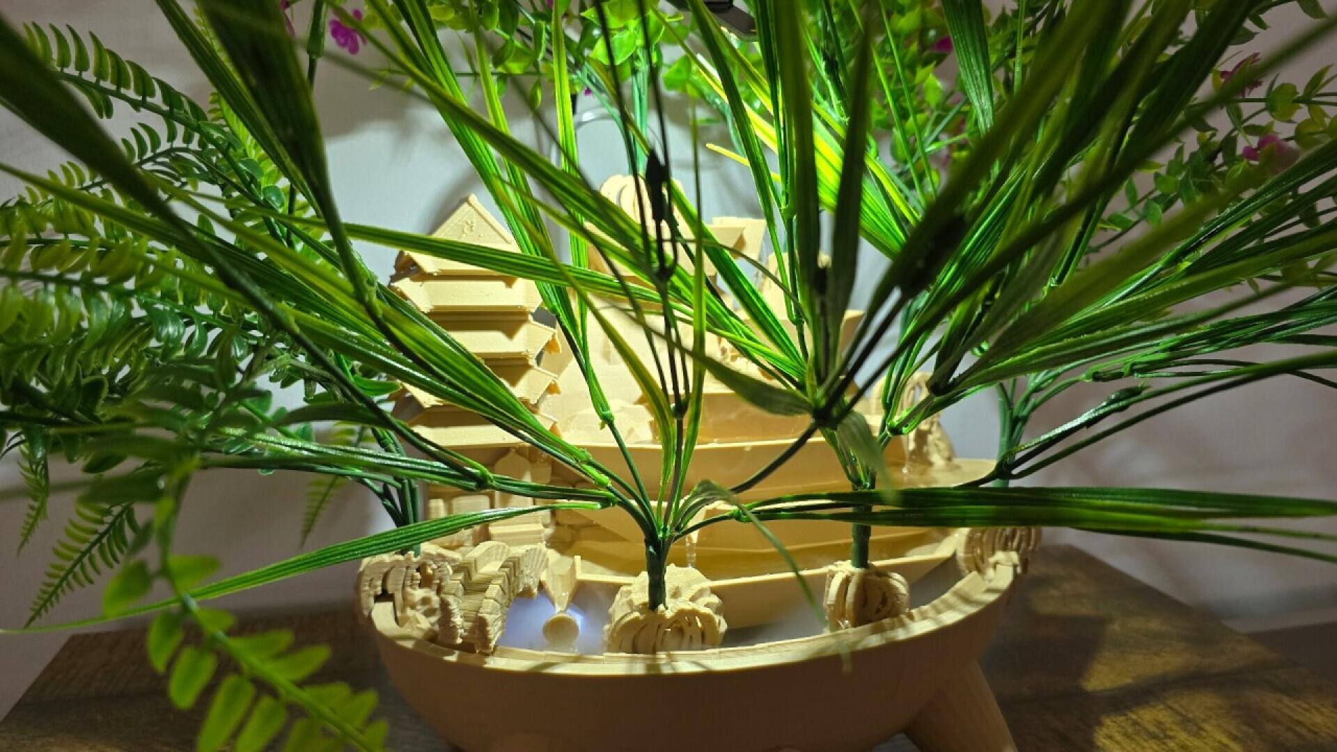 A wooden model of a house is partially hidden behind green artificial plants, which have long, slender leaves. The scene is set on a wooden surface with a white background.