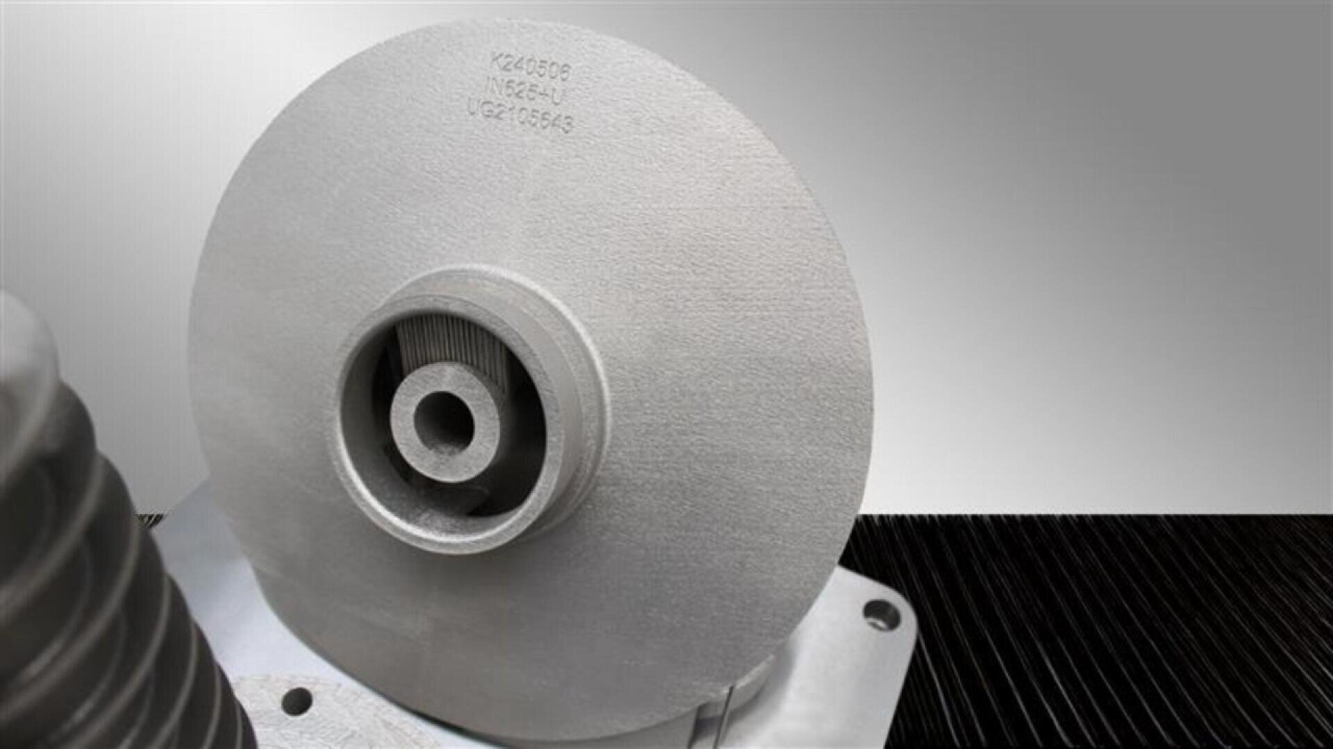 Close-up of a metallic, circular industrial component with a central hole, positioned on a metal base. The background is neutral gray, and numbers are faintly visible on the surface of the disc.