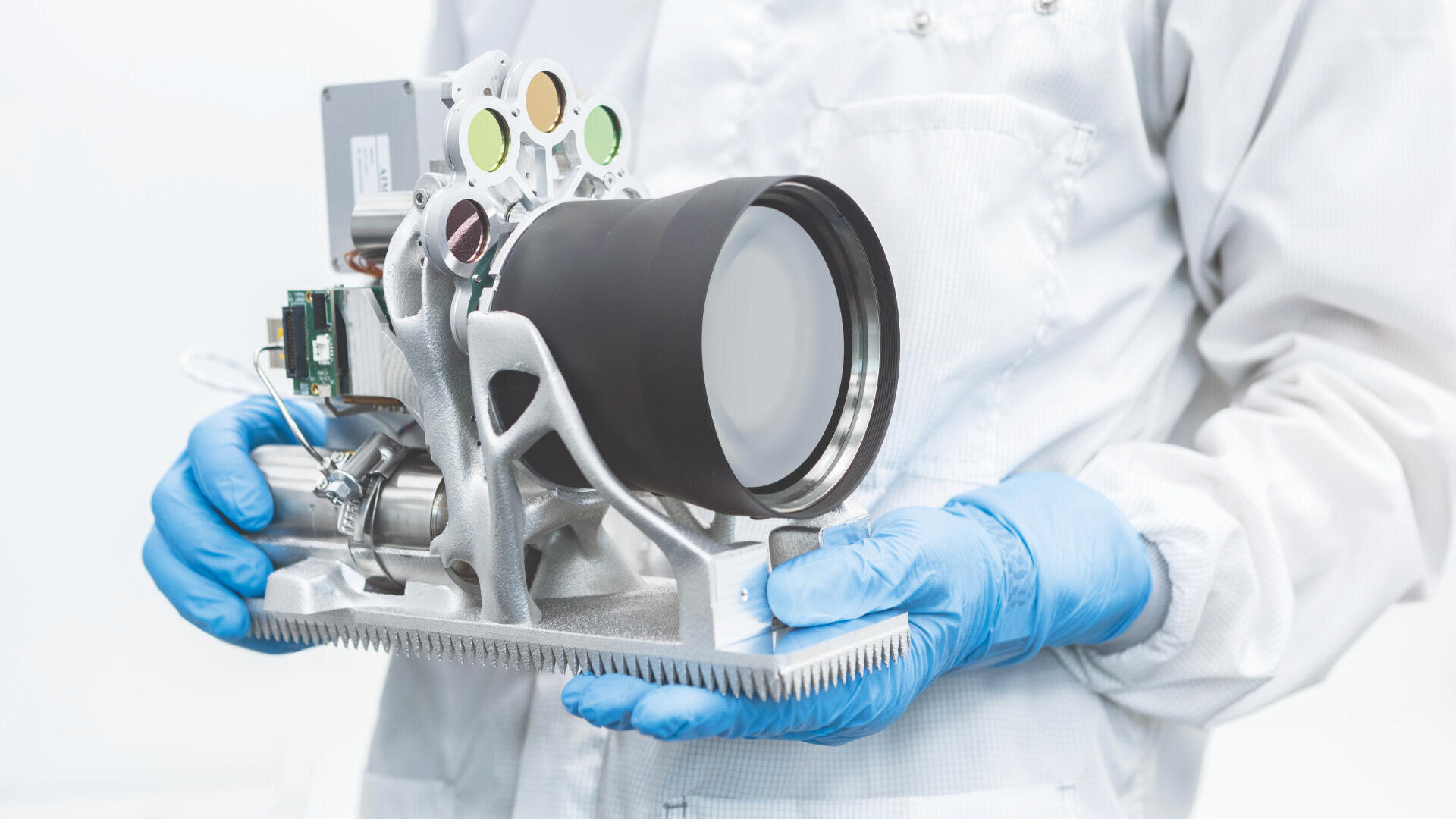 Eine Person in einem weißen Laborkittel und blauen Handschuhen hält vor einem weißen Hintergrund ein komplexes wissenschaftliches Instrument mit einer großen Linse und elektronischen Komponenten.