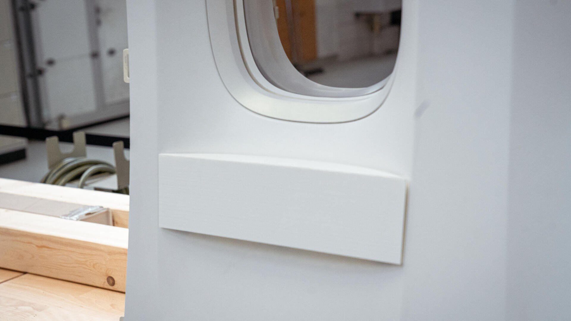 Nahaufnahme eines Flugzeuginnenwandausschnitts mit einem Fenster und einer rechteckigen Platte darunter; links sind ein Teil einer Holzkiste und einige Kabel zu sehen.