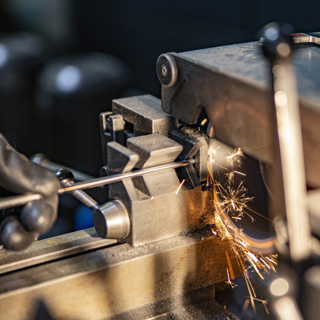 Nahaufnahme einer Metallbearbeitungsmaschine, die einen Metallstab schleift. Sichtbare Funken fliegen. Eine behandschuhte Hand hält den Stab fest gegen die Schleifscheibe.